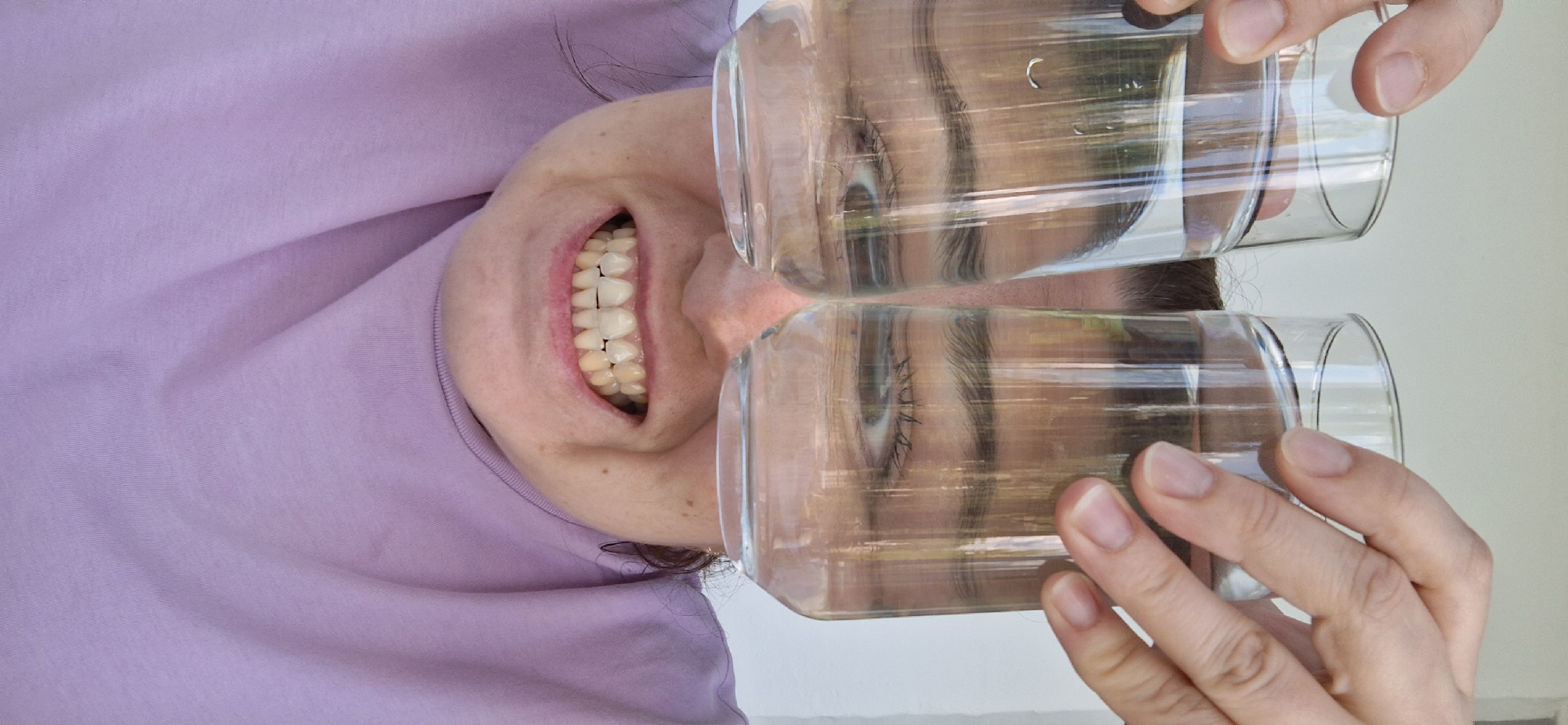 The image depicts a woman holding two clear glasses of water in front of her eyes, creating a distorted view of her face. Her mouth is open, revealing her teeth. She is wearing a lilac-colored shirt. The location is unknown and cannot be determined from the image alone. The city of Chaikovsky, Russia, is not identifiable within the scene.