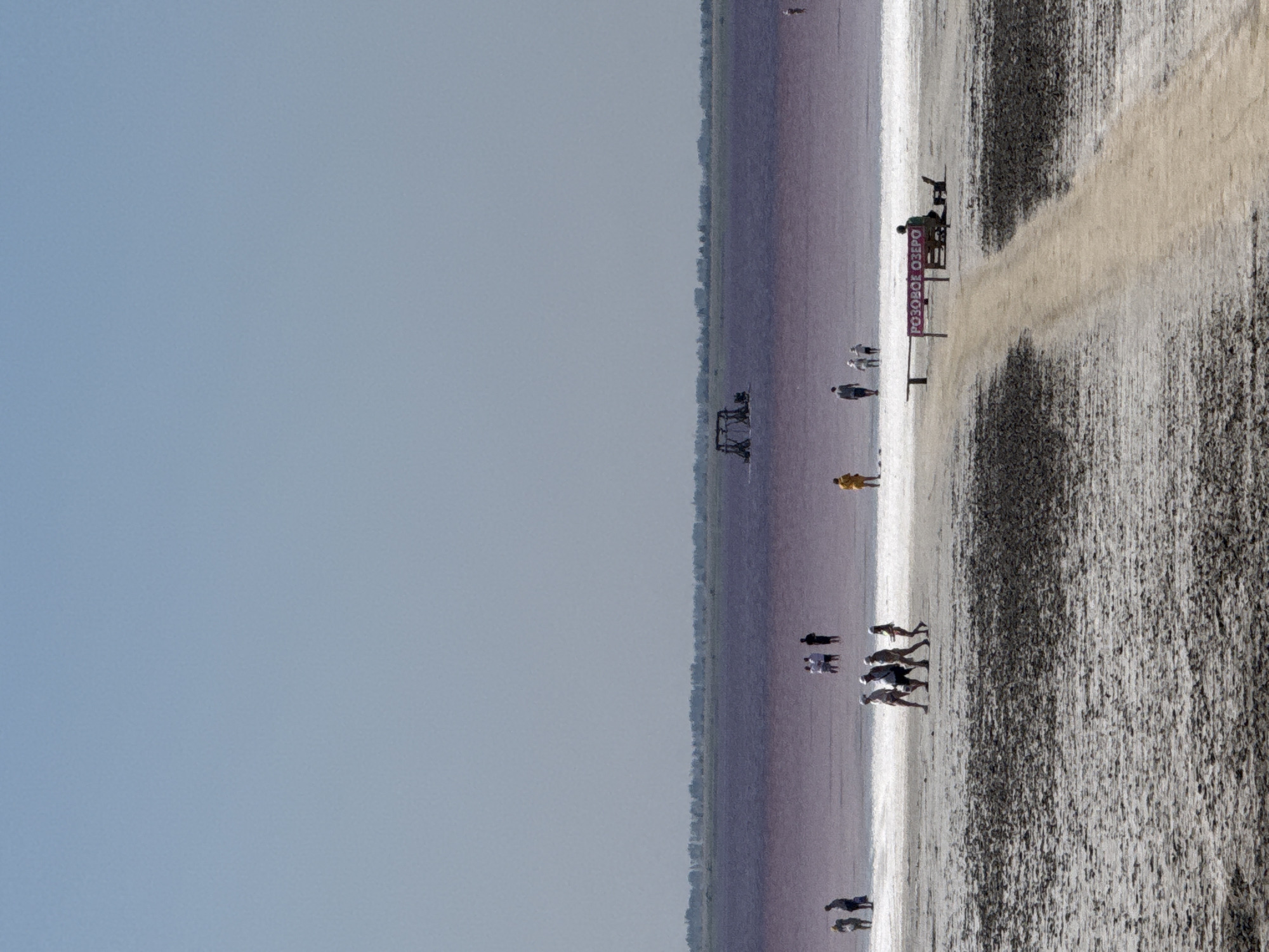 The image depicts a wide, flat, salt-covered landscape with a pink-hued body of water in the distance. The foreground is a dry, cracked, white and dark gray salt flat. A dirt path leads from the foreground towards the water. Several individuals are scattered across the landscape. In the middle ground, on the edge of the salt flats, a group of people are walking or standing near the water. Further into the pink water, a small wooden structure resembling a swing set is visible. In the far distance, a line of trees forms the horizon. A red sign with white Cyrillic text, "РОЗОВОЕ ОЗЕРО" (Pink Lake), is placed near the dirt path, adjacent to a wooden bench and a seated person. The sky is a clear, pale blue. The overall scene suggests a tourist destination focused on the unique pink lake, likely the Boursoľ salt lake in Russia.