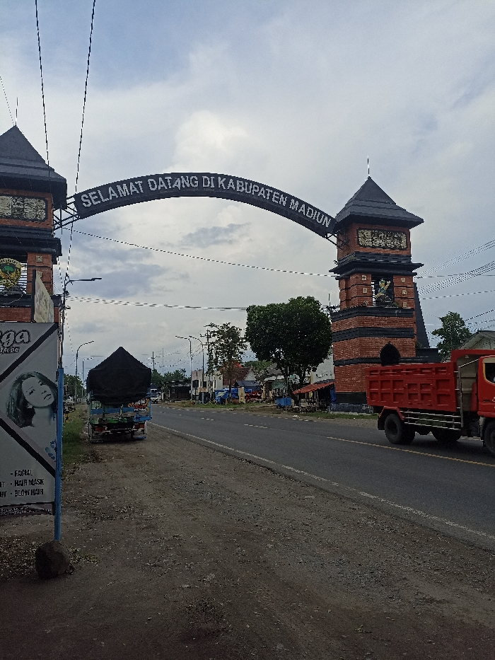 The scene is a road with a large archway in the background. The archway is made of brick and has a black roof. There is a sign on the archway that says "SELAMAT DATANG DI KABUPATEN MADIUN". There is a red dump truck driving on the road. There is a black truck parked on the side of the road. There are some buildings on the side of the road. There is a sign on the side of the road that says "FORMAI - HAIR MASK - BLOW HAIR". There is a woman's face on the sign. There are trees on the side of the road. The sky is overcast with clouds. This scene is in Kecamatan Pilangkenceng, Indonesia. The coordinates are -7.492 latitude and 111.604 longitude.