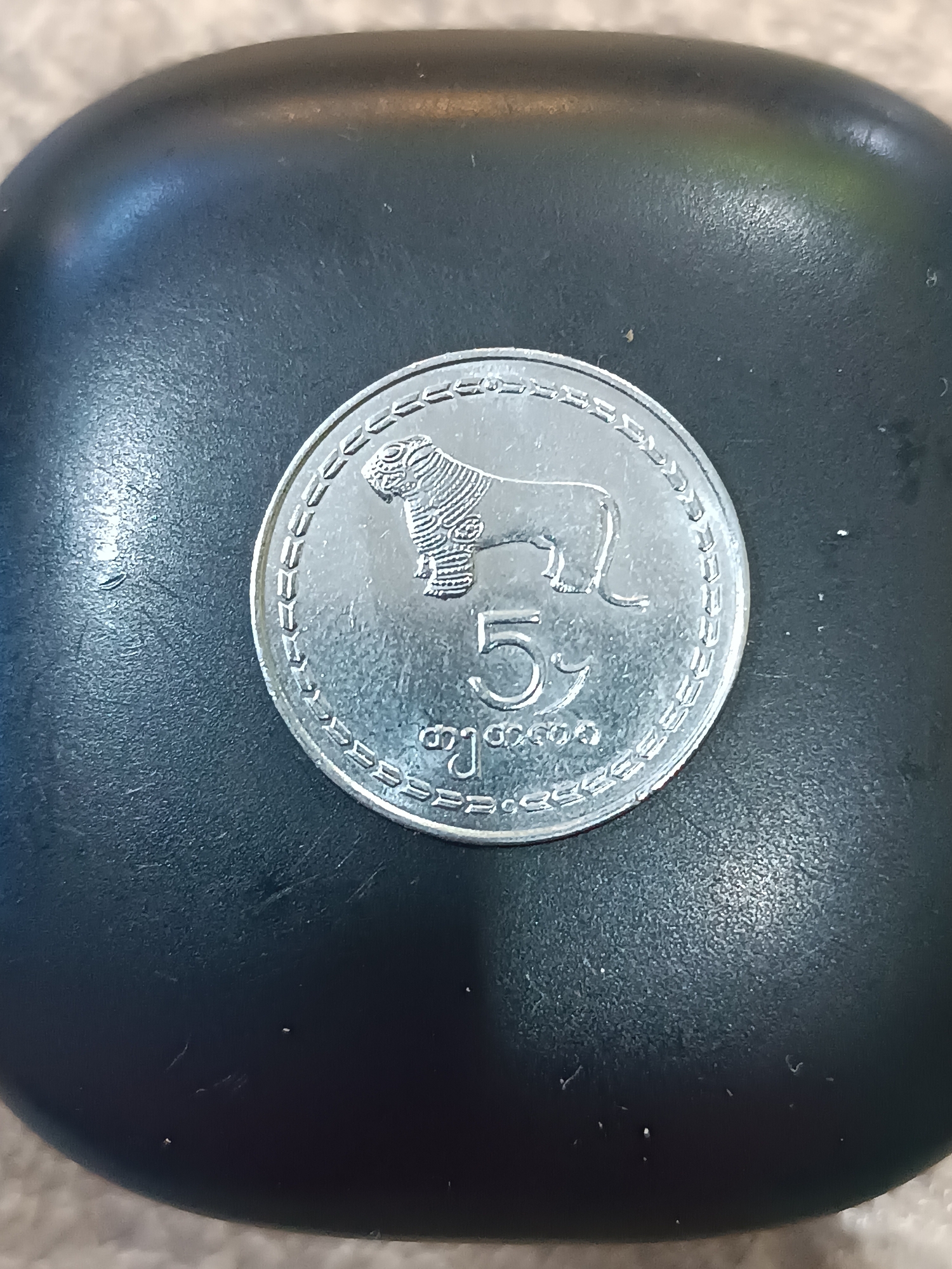 A silver coin featuring a lion and the number "5" is placed on a dark, textured surface. The coin has a decorative border. The surface appears to be a rounded object, possibly a device or an accessory. The lighting suggests an indoor setting. There is no visible indication of the coin's location in Khashuri, Georgia.