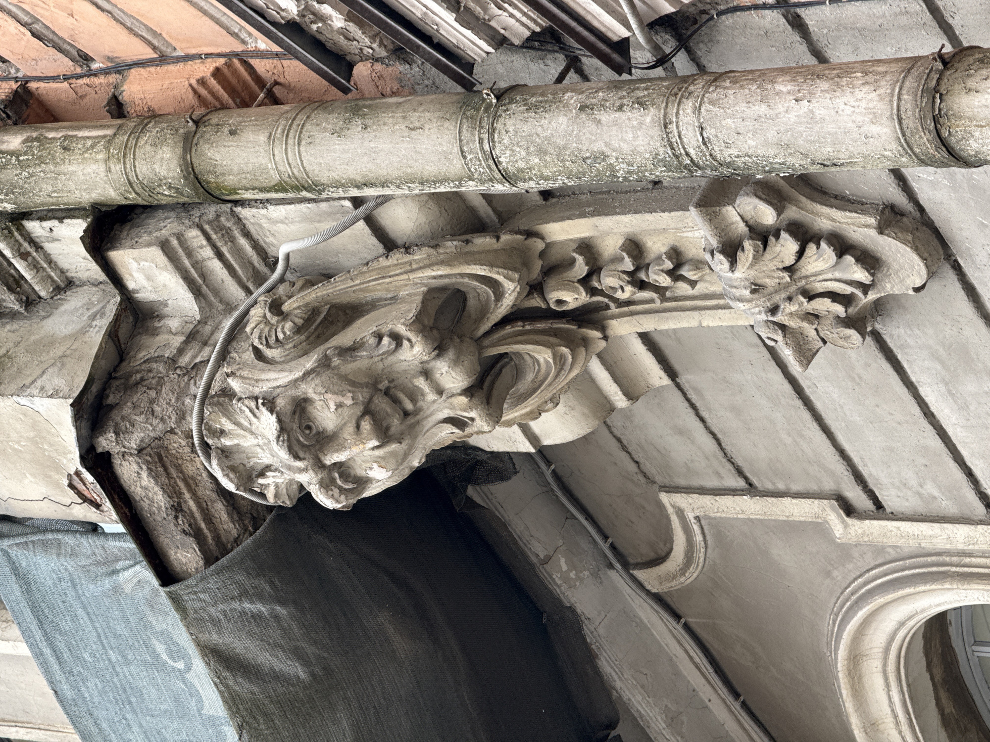 The image shows a close-up, eye-level view of architectural details on the exterior of an old building in Saint Petersburg, Russia. The focal point is a weathered stone grotesque face protruding from the facade, serving as a corbel. The grotesque has deeply carved features, including wide eyes, a prominent nose, and an open mouth, with decorative elements resembling draped fabric or hair flowing downwards. Below the face, there is a carved floral motif.
Adjacent to the grotesque, a vertical, grey downspout runs down the side of the building. The downspout is cylindrical with ribbed sections and shows signs of age and wear, including peeling paint and moss or discoloration. A metal clamp secures it to the wall.
The building's facade is made of aged plaster, with visible cracks and signs of decay, particularly around the upper section where a torn grey fabric or tarp is draped over a damaged architectural element. The tarp is secured by what appears to be netting. To the right of the downspout, a section of brick wall in a reddish-orange hue is visible, with some electrical conduits and wires running along it.
In the lower left portion of the image, a curved window frame with intricate detailing is partially visible, suggesting the presence of an arched window. The overall impression is one of historical architecture in a state of disrepair, possibly undergoing renovation or simply showing the effects of time and weather. The lighting is even, indicating an overcast day or shade.