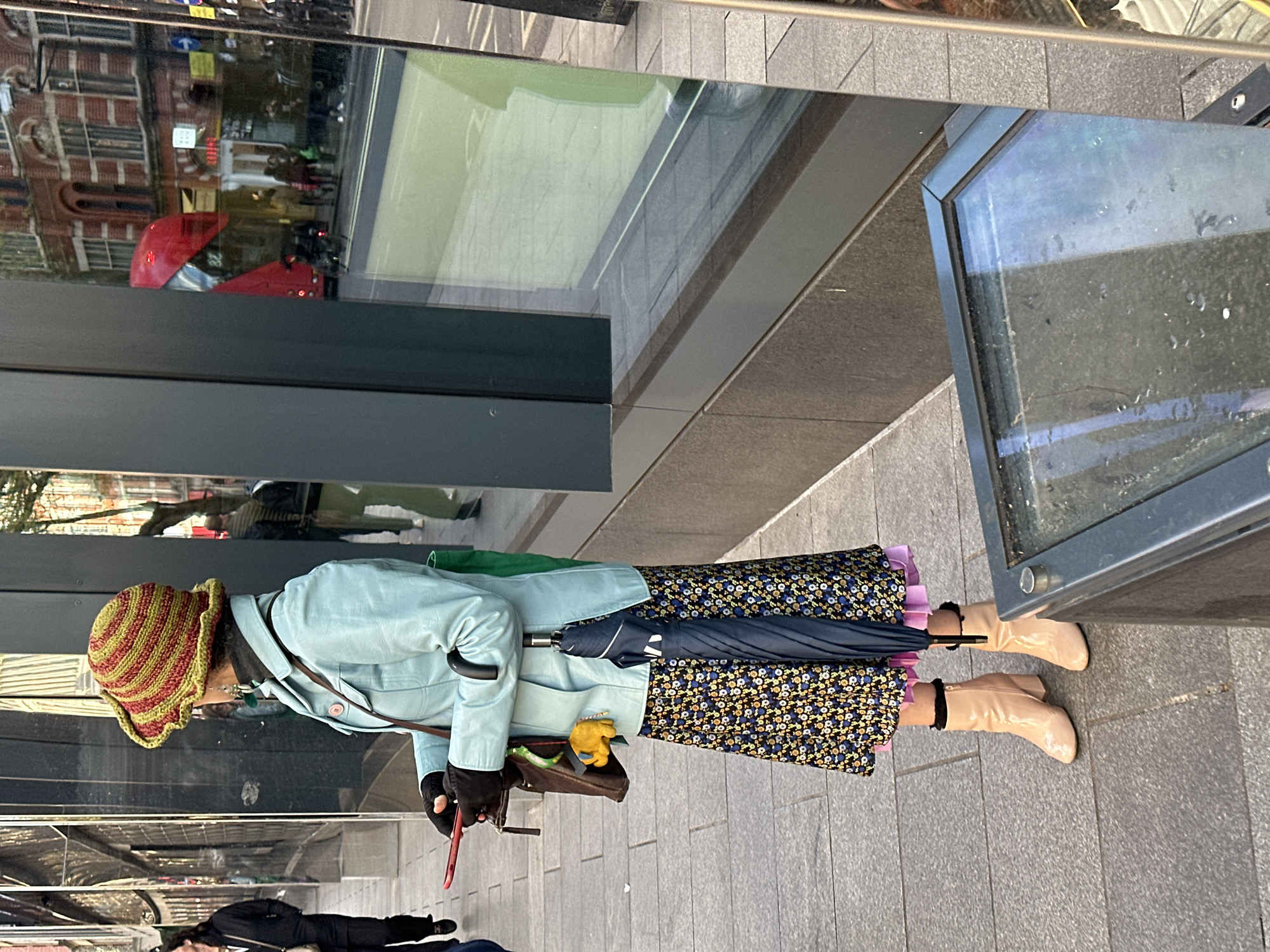 An adult female stands on a paved sidewalk in London, United Kingdom. She wears a red and yellow striped hat, a light blue jacket, a floral patterned skirt, and beige boots. She holds a closed, dark-colored umbrella and a red mobile phone or similar device. Beside her is a building with mirrored windows reflecting the urban environment. To the right of her is what appears to be an advertisement stand or an information kiosk. Other people are walking past in the background.