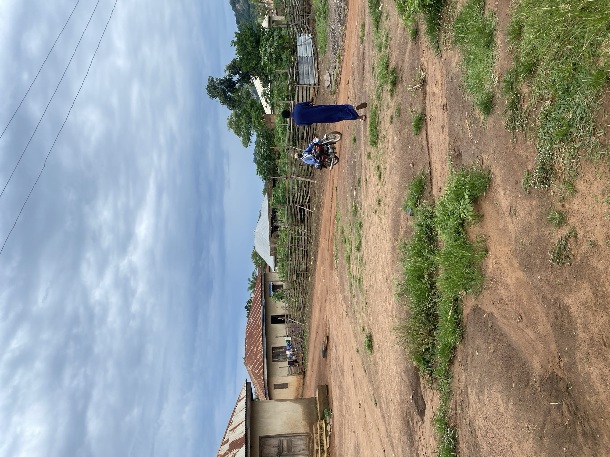 A medium shot shows a dirt road with a man in a long blue robe walking away from the camera. To his left, a person on a red motorcycle is riding towards the camera. The man in blue is walking away from a village with several small houses with corrugated metal roofs. The houses are made of earth and cement. A wooden fence runs along the edge of the village. The sky is overcast with clouds. The scene suggests a rural setting in a warm climate.