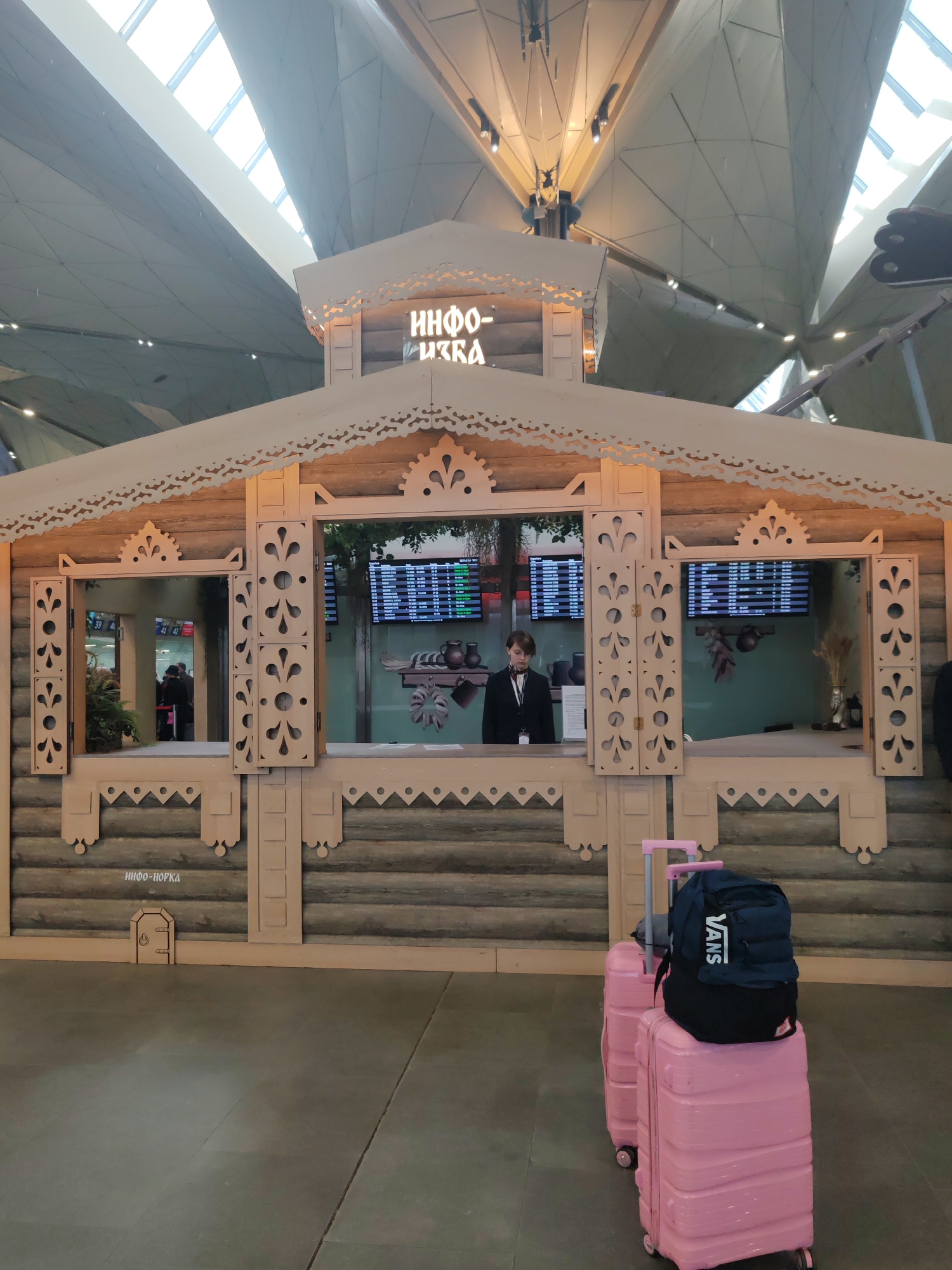 This full-shot image captures a unique information booth, designed to resemble a traditional Russian log house (izba) with ornate wooden carvings, set within a modern, spacious interior. The high, angular ceiling features large skylights and exposed structural elements, indicating a contemporary architectural style typical of a major airport terminal.
At the center of the wooden booth, a person, likely a female attendant, dressed in a dark uniform with a light shirt and dark tie, stands behind a counter, facing forward. Behind them, digital screens display what appears to be flight or schedule information, along with traditional pottery and dried decorations on shelves. The booth's sign, in Cyrillic, prominently reads "ИНФО-ИЗБА" (INFO-IZBA), emphasizing its traditional Russian theme. The same text is faintly visible on the dark, tiled floor in front of the booth.
In the foreground to the right, two bright pink hardshell suitcases are stacked, with a dark blue backpack, marked "VANS" in white lettering, resting on top of the front suitcase. The presence of luggage and the information screens strongly suggest an airport setting.
The scene takes place during the daytime, evidenced by natural light filtering through the skylights. The traditional design of the information booth creates a striking contrast with the sleek, modern architecture of the surrounding environment. This blend of old and new, along with the Cyrillic text and traditional architectural elements, is consistent with a location such as Pulkovo Airport in Saint Petersburg, Russia.