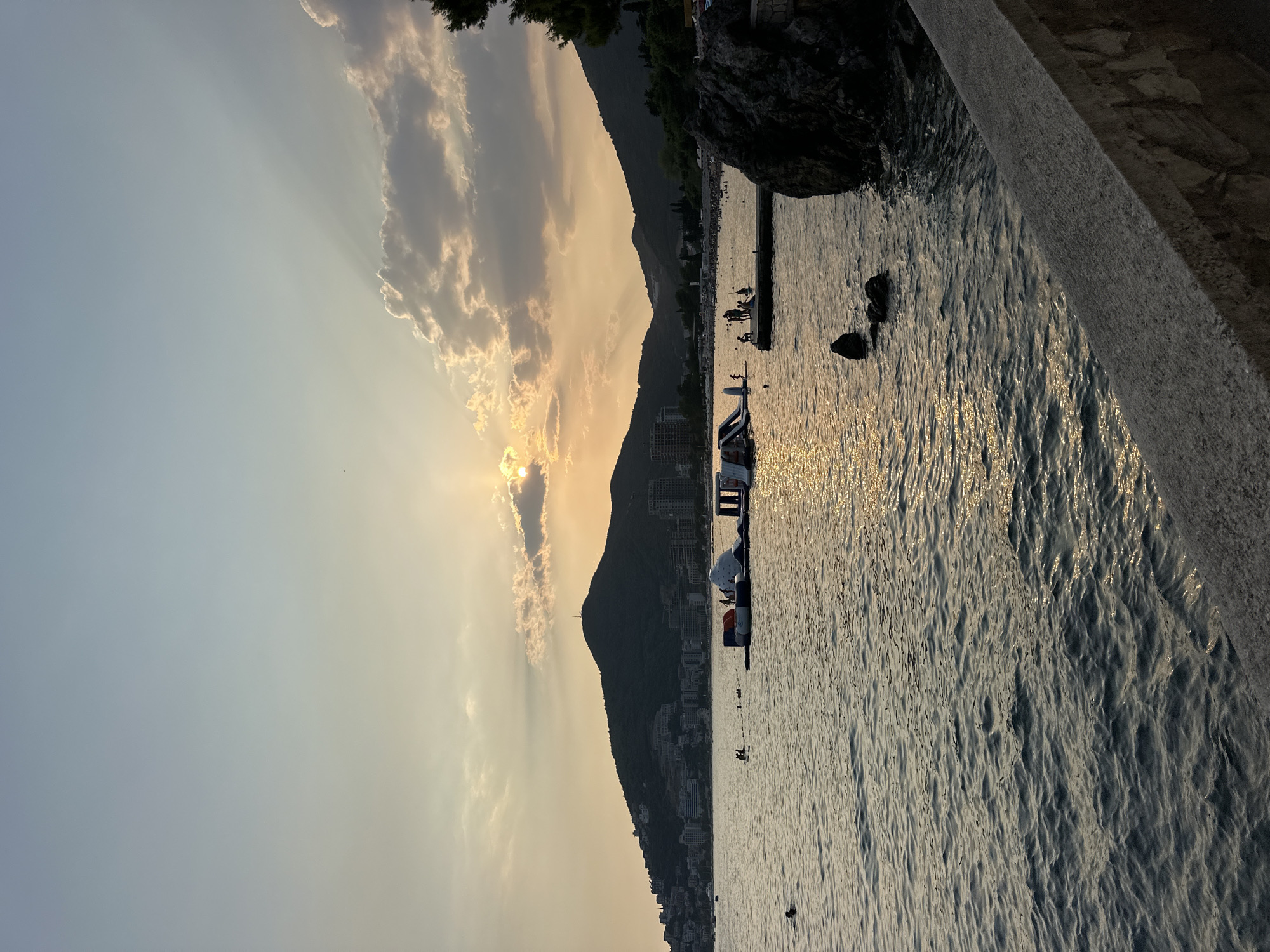 The image depicts a sunset scene at Mogren Beach in Budva, Montenegro. The sun is setting behind a mountain range that overlooks the coastline, with its rays filtering through scattered clouds. The water is calm with gentle waves reflecting the warm light. In the foreground, a stone embankment lines the edge of the water.
In the middle ground, an inflatable water park with slides and various structures is visible in the sea. Several people are engaged in activities on and around the water park, including some swimming and standing on a wooden platform connected to it. Further out, other individuals can be seen swimming in the sea.
The background shows a hillside densely populated with buildings, characteristic of the coastal town of Budva, nestled against the mountains. The overall atmosphere is serene, with the golden hour light creating a picturesque view of the Adriatic Sea.