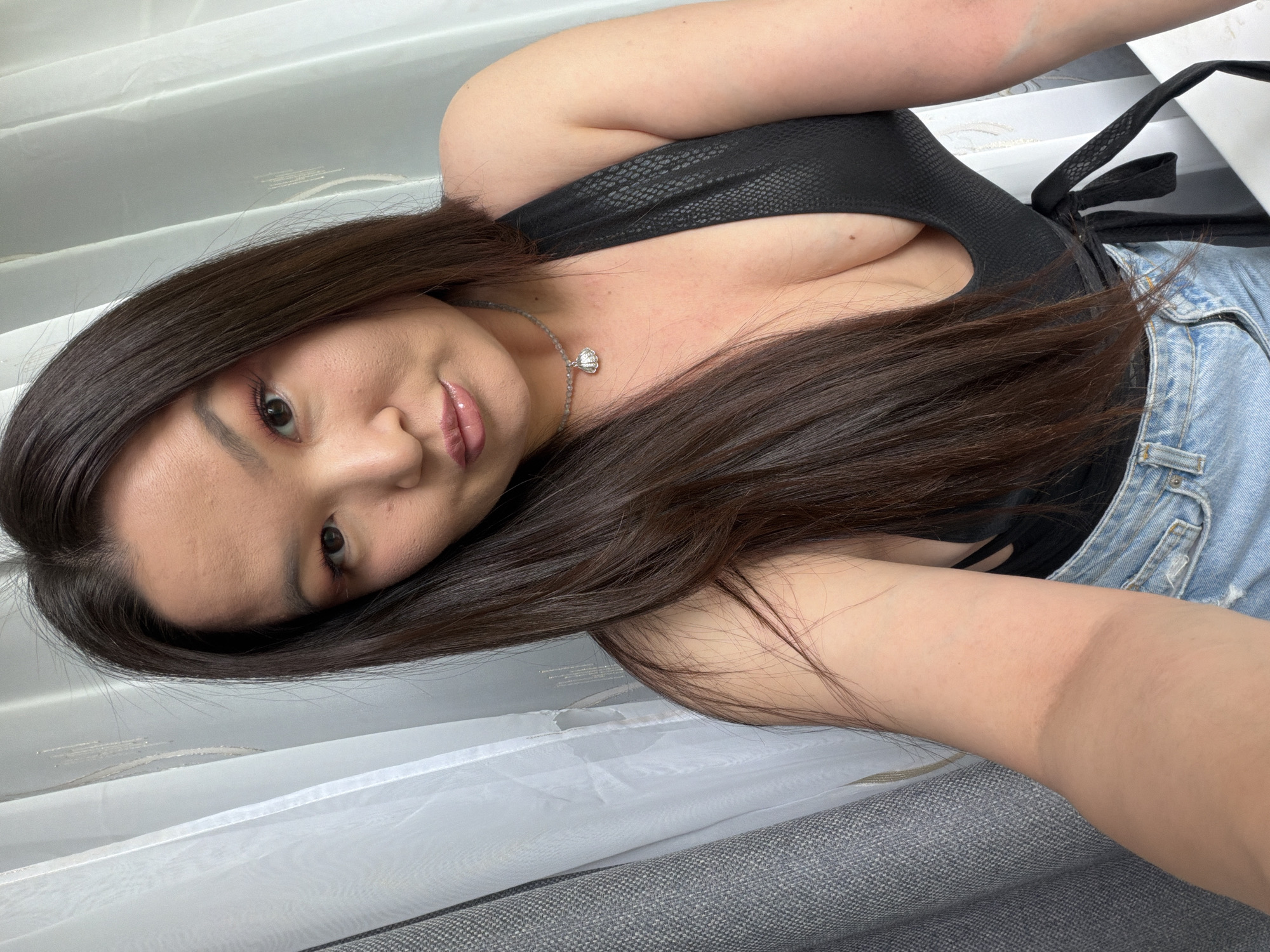 A young woman is depicted in a selfie-style image. She has long, dark hair that cascades over her left shoulder and is partially obscuring her chest. She is wearing a black top with a textured pattern and a pair of light blue denim shorts. A silver necklace with a pendant is visible around her neck. Her makeup includes a light shade of eyeshadow and glossy lips. The background features a white curtain that is partially visible behind her. The scene does not contain enough information to determine the location in any city.
