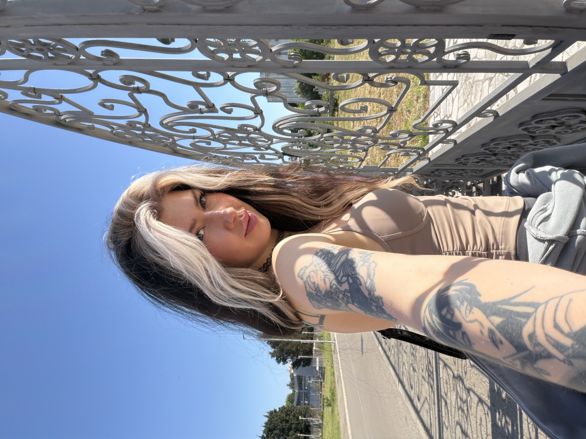 A woman is posing for a selfie in Šušanj, Montenegro. She has long brown hair with a streak of blonde, fair skin, and visible tattoos on her arms. She is wearing a beige top and a grey jacket. The background features a wrought-iron fence, a grassy area, a building, a road, and a clear blue sky.