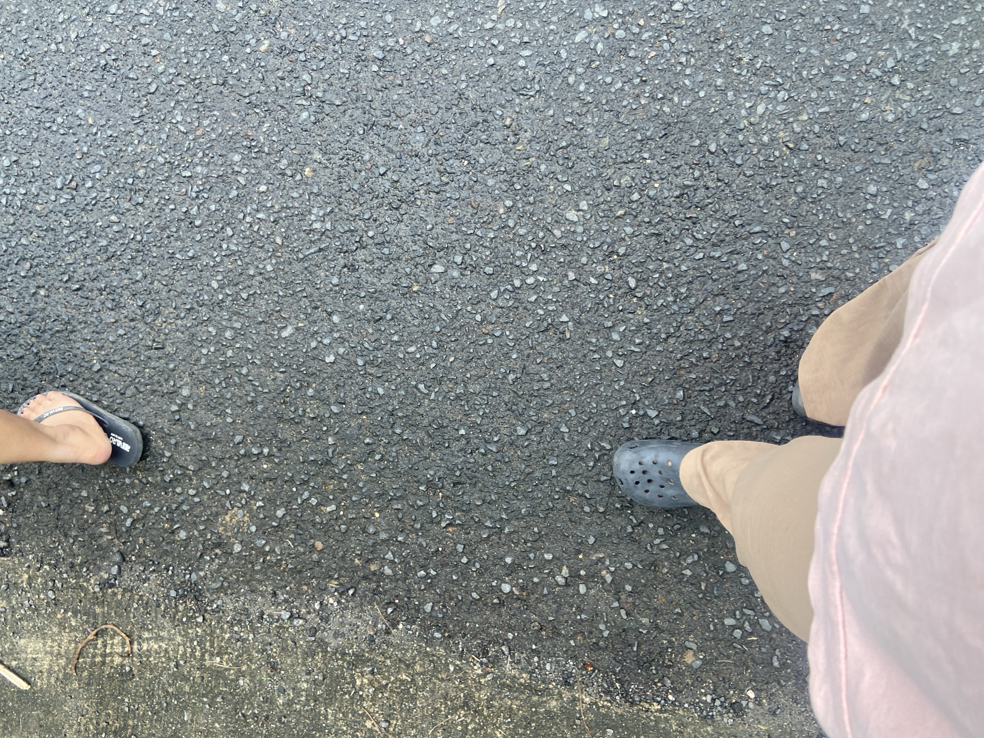 A first-person perspective shot shows two feet on a dark asphalt surface. The left foot is wearing a dark blue Croc-style shoe. The right foot, positioned higher in the frame, is wearing a black flip-flop, revealing a painted toenail. The person's legs are visible, with the left leg clad in light brown pants and the right leg in a light pink garment that appears to be a skirt or dress. The asphalt surface is speckled with small grey stones. To the left of the frame, a section of lighter, possibly concrete or dirt, ground is visible with a small, curved brown twig lying on it. The image captures a simple scene of walking on a paved surface, possibly a street or sidewalk in Makale, Indonesia.