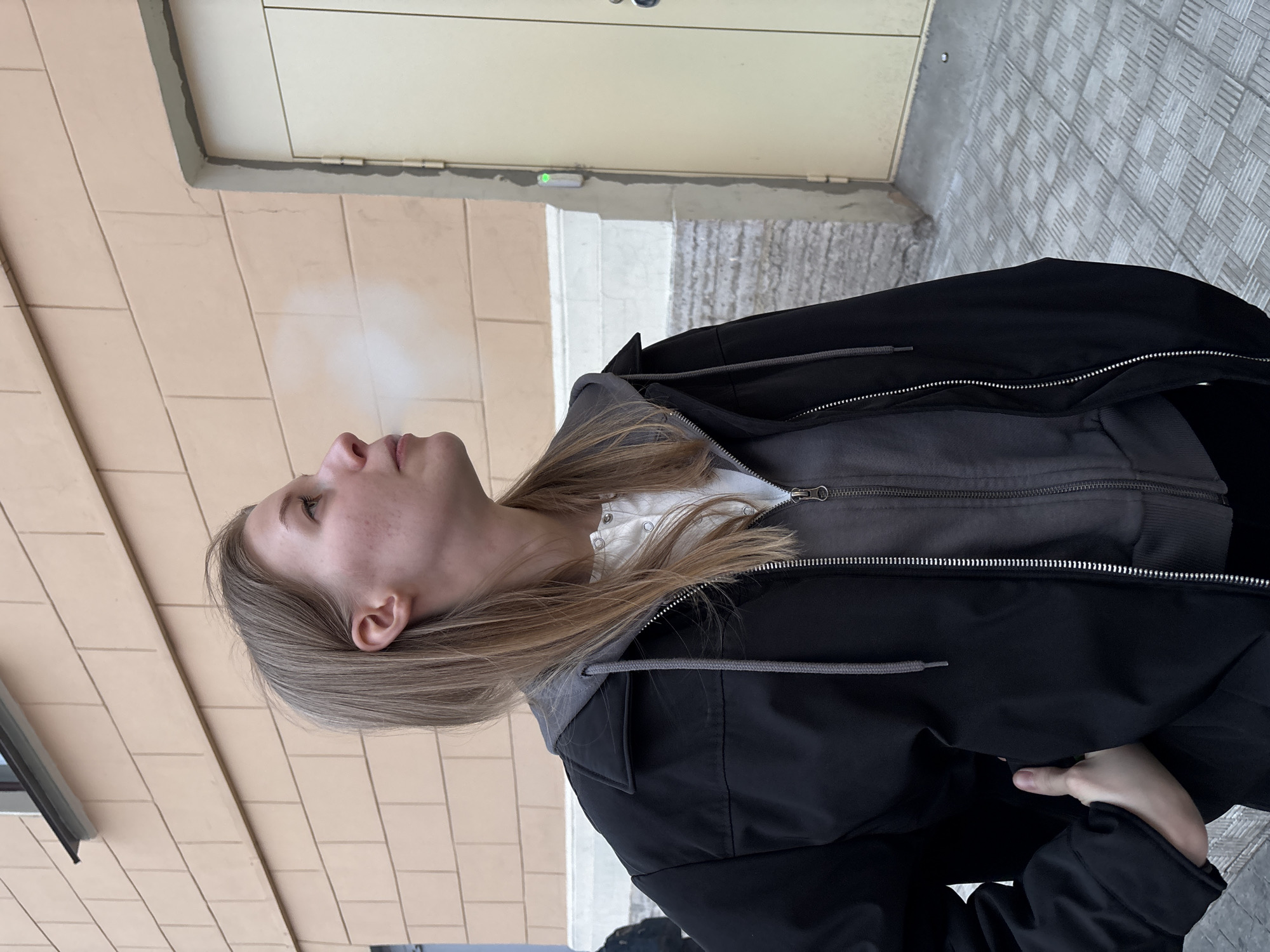 A young, light-skinned female stands outdoors, exhaling vapor that is visible in the cold air. She has long blonde hair, a closed mouth, and is looking upwards. She is wearing a white, buttoned shirt with a gray hooded sweatshirt, covered by a black jacket with a zipper. Her left hand is visible in the lower right corner. The background consists of an exterior wall of a building in shades of tan and beige, and what appears to be a sidewalk with textured tiles. The setting is identified as Saint Petersburg, Russia based on architectural and ambient clues.