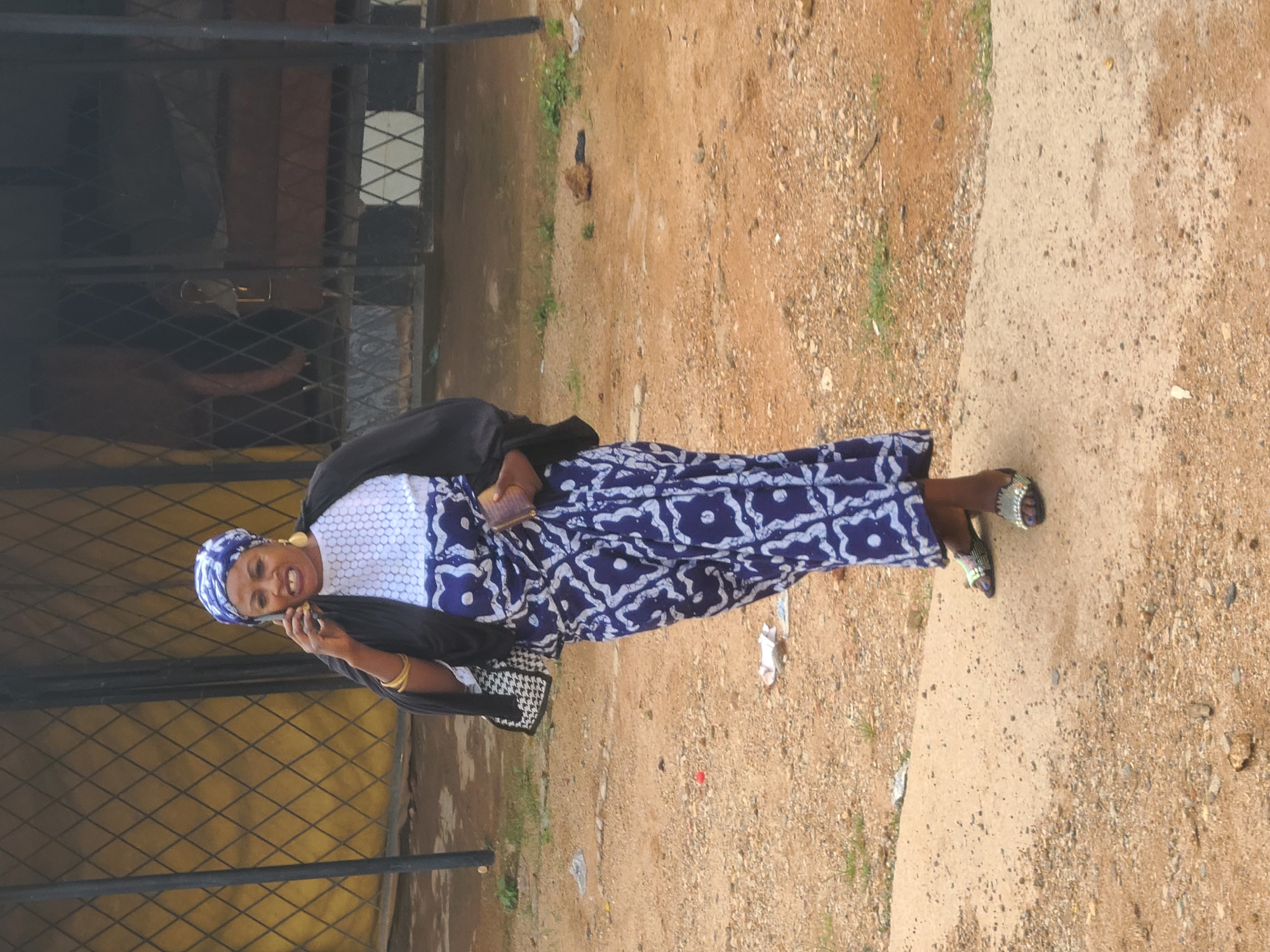 A woman is standing outdoors, seemingly on a paved area adjacent to a dirt path or ground. She is dressed in a long, patterned blue and white dress, a white top underneath, and a black jacket. She has a matching blue and white headwrap, gold earrings, and gold bracelets. The woman is holding a mobile phone to her ear, suggesting she is engaged in a conversation. Behind her is a yellow metal gate. The surrounding environment includes a dirt path with sparse vegetation. The location is in the city of Unknown, Unknown.