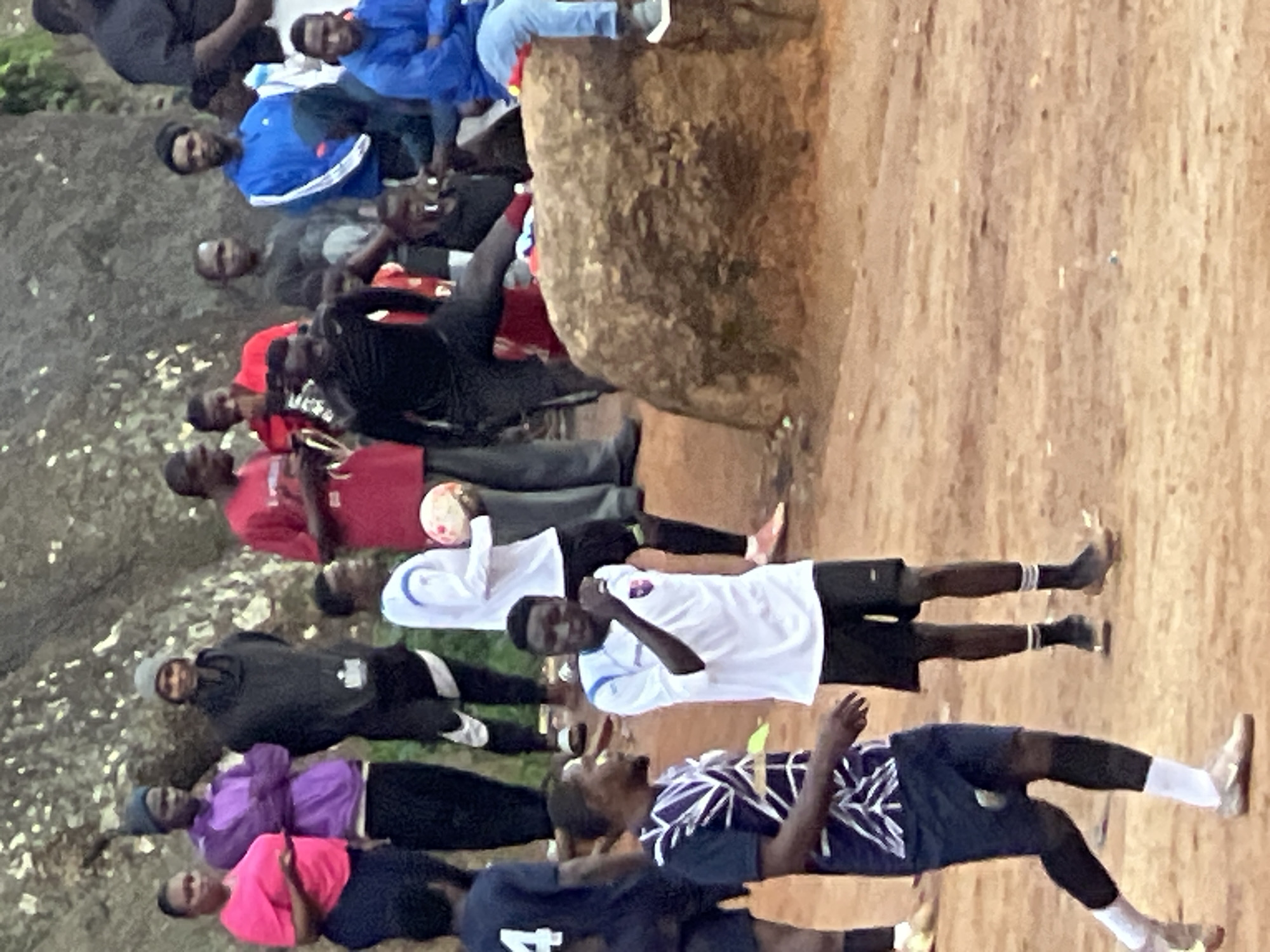 In the city of Jos, Nigeria, a group of young men are gathered outdoors, possibly for a soccer match. The ground is a reddish-brown dirt, and a large, rough-textured rock formation dominates the background. Several individuals are dressed in athletic wear, with some in soccer jerseys and shorts, while others wear casual clothing like hoodies and t-shirts. One person is holding a soccer ball. The atmosphere appears to be informal, with people standing and observing. The lighting suggests it is daytime, with soft, diffused light.