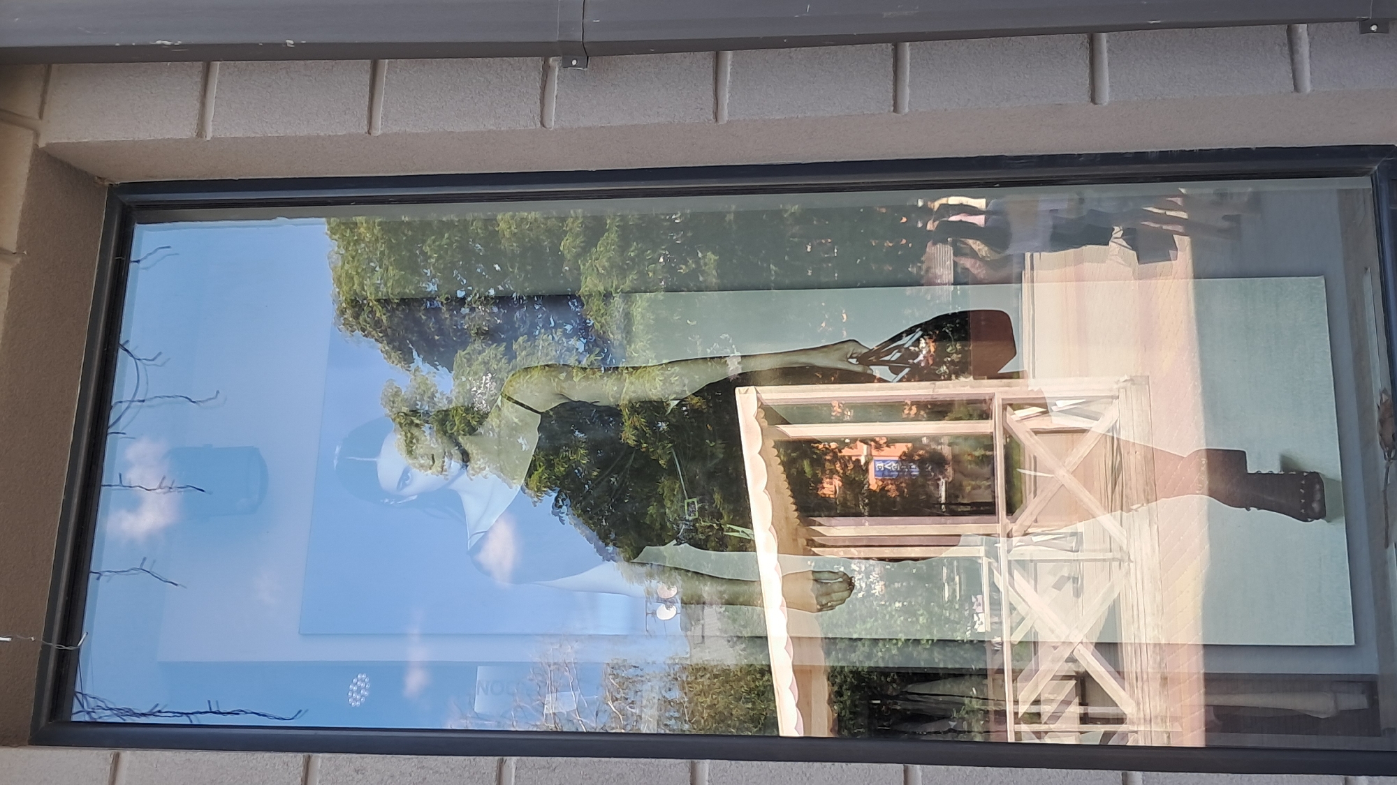 A shop window displays a large print of a woman wearing a black slip dress and high-heeled boots. She is holding a handbag. The window reflects the sky with clouds, trees, and buildings. The shop is located on Ternopil, Ukraine.