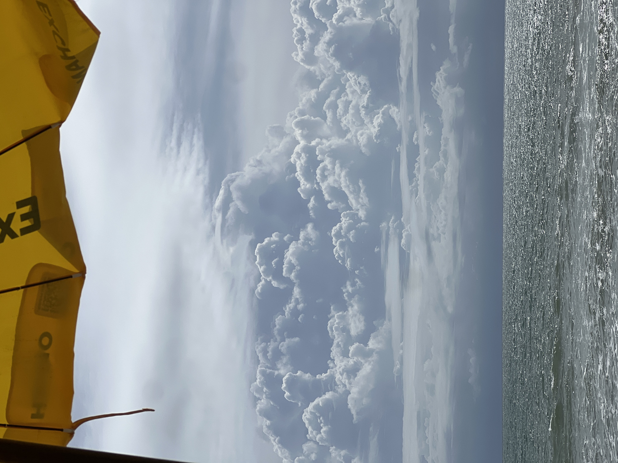 The image presents a wide view of an ocean and sky from a low perspective, framed by the underside of a yellow umbrella. The umbrella occupies the top portion of the frame, displaying reversed dark lettering, including "EXCH" and "OH" or "HO", along with part of a support pole on the left and a hanging reddish-brown strap. The sky is heavily clouded, featuring a prominent, voluminous formation of bright white and grey cumulus clouds dominating the upper mid-ground, with flatter, darker stratiform clouds above and below. The lower half of the image shows the ocean, its surface appearing dark grey-green, choppy, and shimmering with reflected light, particularly on the waves in the immediate foreground where some white foam is visible. A clear horizon line separates the ocean from the dense cloud base. There are no individuals or identifiable man-made structures beyond the umbrella. The location is in the city of Unknown, Unknown.