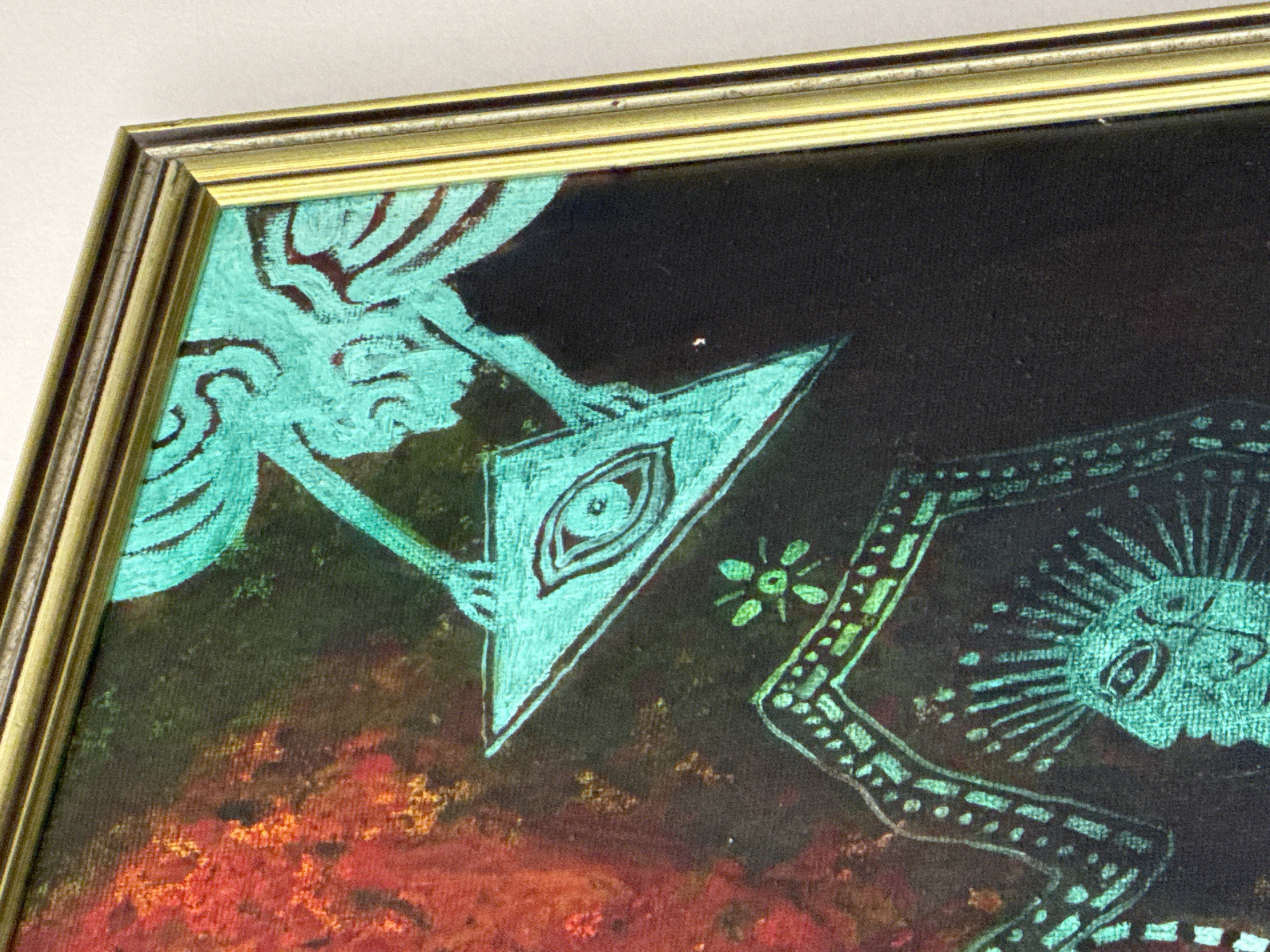 A framed painting featuring surreal imagery is presented at an angle. The dominant colors are black, red, and turquoise. In the upper left, a stylized, turquoise figure appears to be holding a turquoise triangle with an eye in the center. To the right of this, there is a small turquoise flower-like motif. Further right, a turquoise pattern resembling a face with radiating lines is situated. The background transitions from black in the upper part to red in the lower part, creating a contrast. The frame has a gold finish. The specific location of Plovdiv, Bulgaria is not discernible in the image.