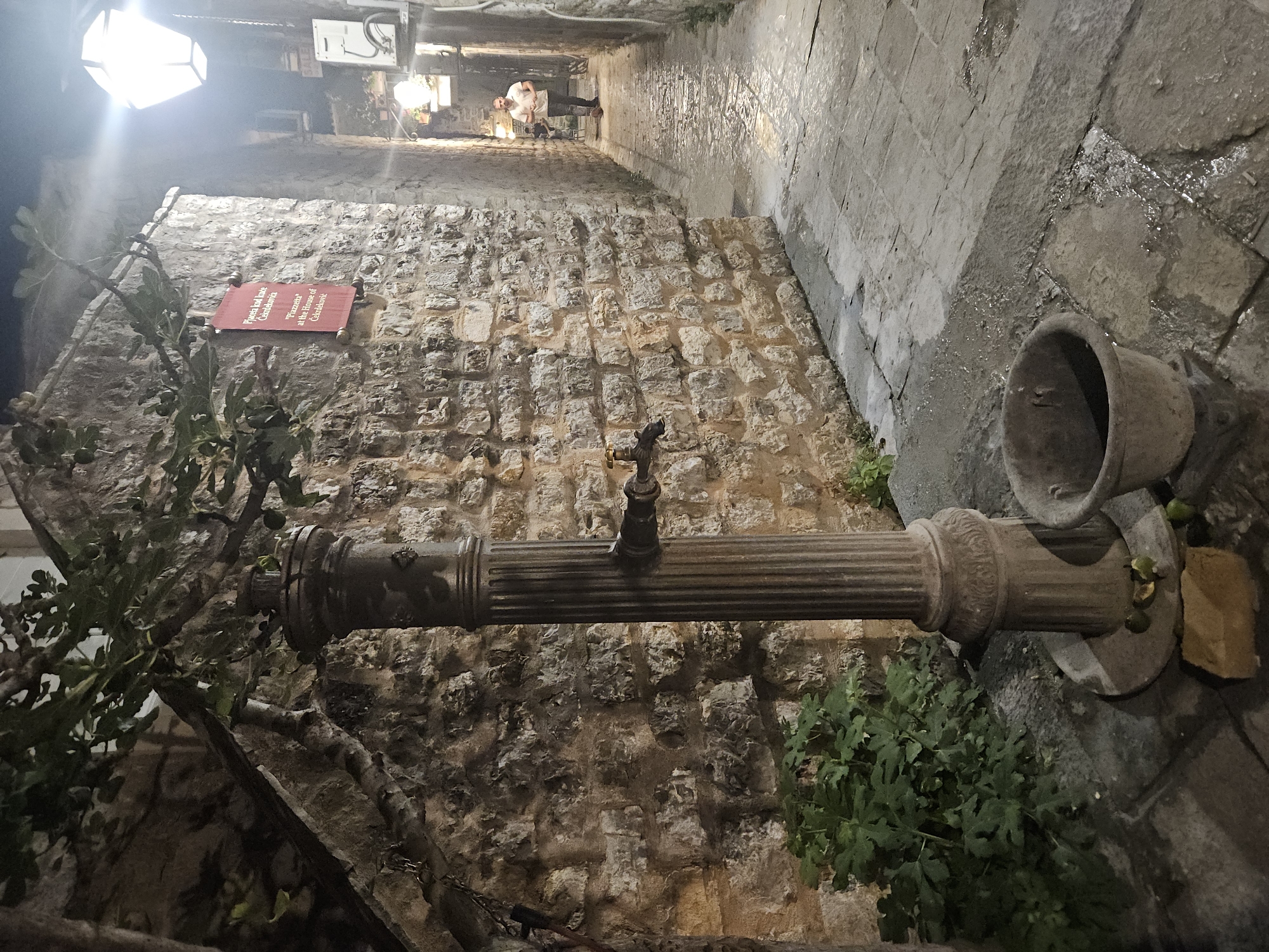 Captured at night, the image showcases a street scene in Unknown, Unknown. A stone water fountain stands in the foreground, featuring a horizontal pipe with a faucet and a bowl to collect water. The background is a stone wall with a red sign bearing white text, and a narrow street lined with stone structures leads into the distance where people and light sources are visible. The stone pavement is wet, suggesting recent rainfall. Foliage grows near the fountain, adding a natural element to the built environment.