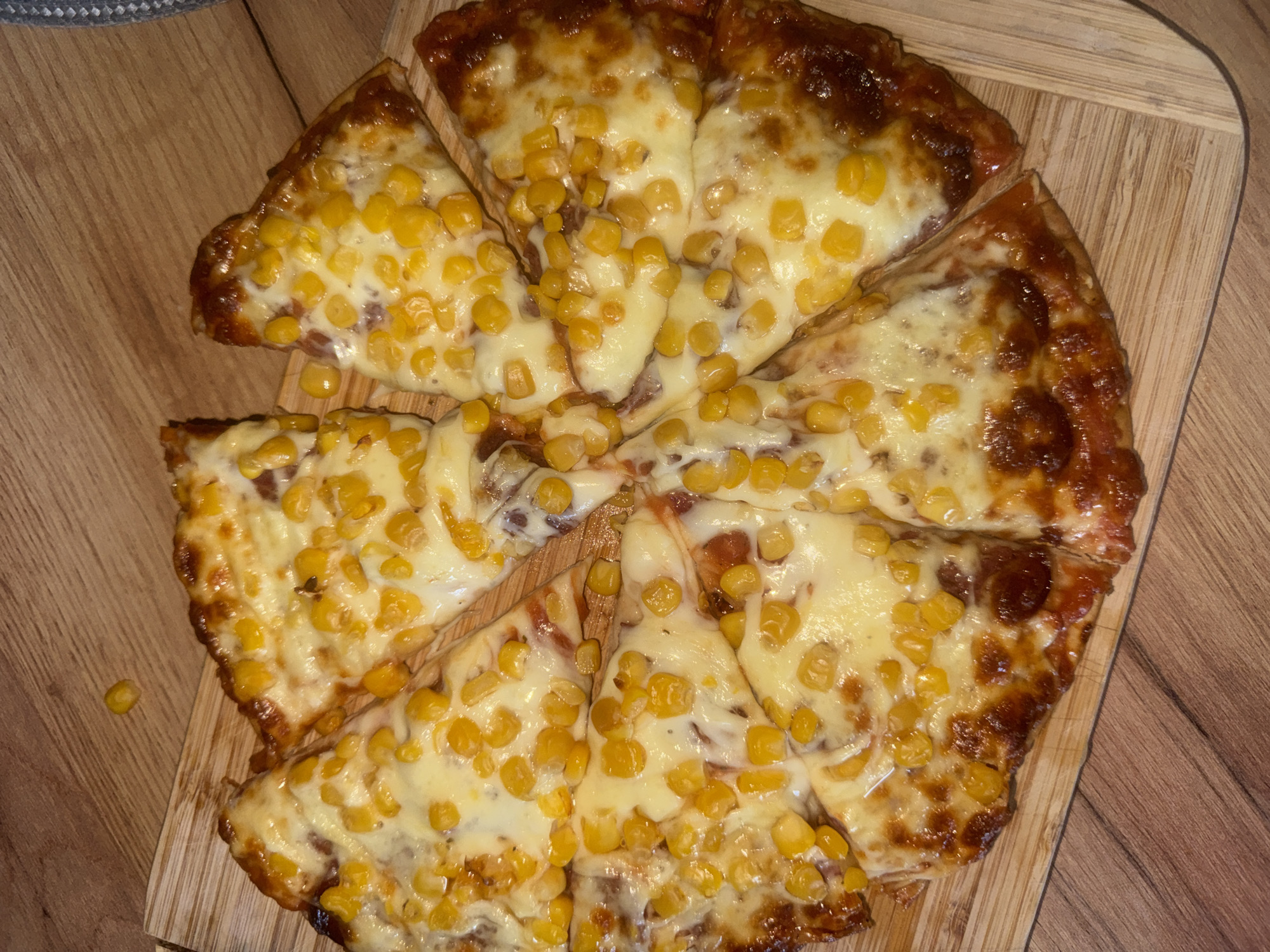A whole pizza, cut into eight slices, sits on a wooden cutting board. The pizza has a red sauce base, melted white cheese, and is topped with corn kernels. The crust is golden brown and slightly charred in places. The cutting board is placed on a wooden table. The image appears to have been taken indoors, possibly in a kitchen or dining area, in the city of Varna, Bulgaria. There are no people visible in the image, and no actions are being performed.