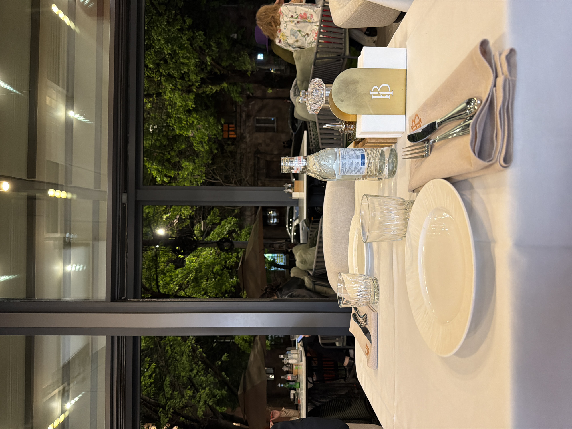 The image captures a set dining table at an upscale restaurant or cafe in Yerevan, Armenia, seen from a guest's perspective, likely at night. The foreground features a neatly arranged place setting on a white tablecloth. A pristine white dinner plate with a subtle textured rim, two clear, cut-glass tumblers, and a small, clear glass bottle of water with a partially obscured label (possibly starting with "Sar...") are visible.
A notable detail is an elegant decanter, possibly for oil or vinegar, with a crystal stopper, positioned on a sleek gold-colored box. This box, along with a beige fabric napkin holder containing a silver-colored knife and fork, prominently displays a stylized "B" or "IB" monogram, suggesting the establishment's branding.
Beyond the table, a large glass partition or window offers a view of the outside. Lush green trees, artificially lit from below, dominate the mid-ground, indicating an evening setting. Reflections of interior lights are visible on the glass. In the background, other patrons are seated at different tables; one person with long, curly brown hair wearing a floral top is visible with their back to the camera. Faintly, a building with some illuminated windows can be seen through the foliage. The overall ambiance suggests a sophisticated dining experience in a well-lit, possibly outdoor or semi-outdoor, setting. No specific activity beyond dining is clearly depicted.