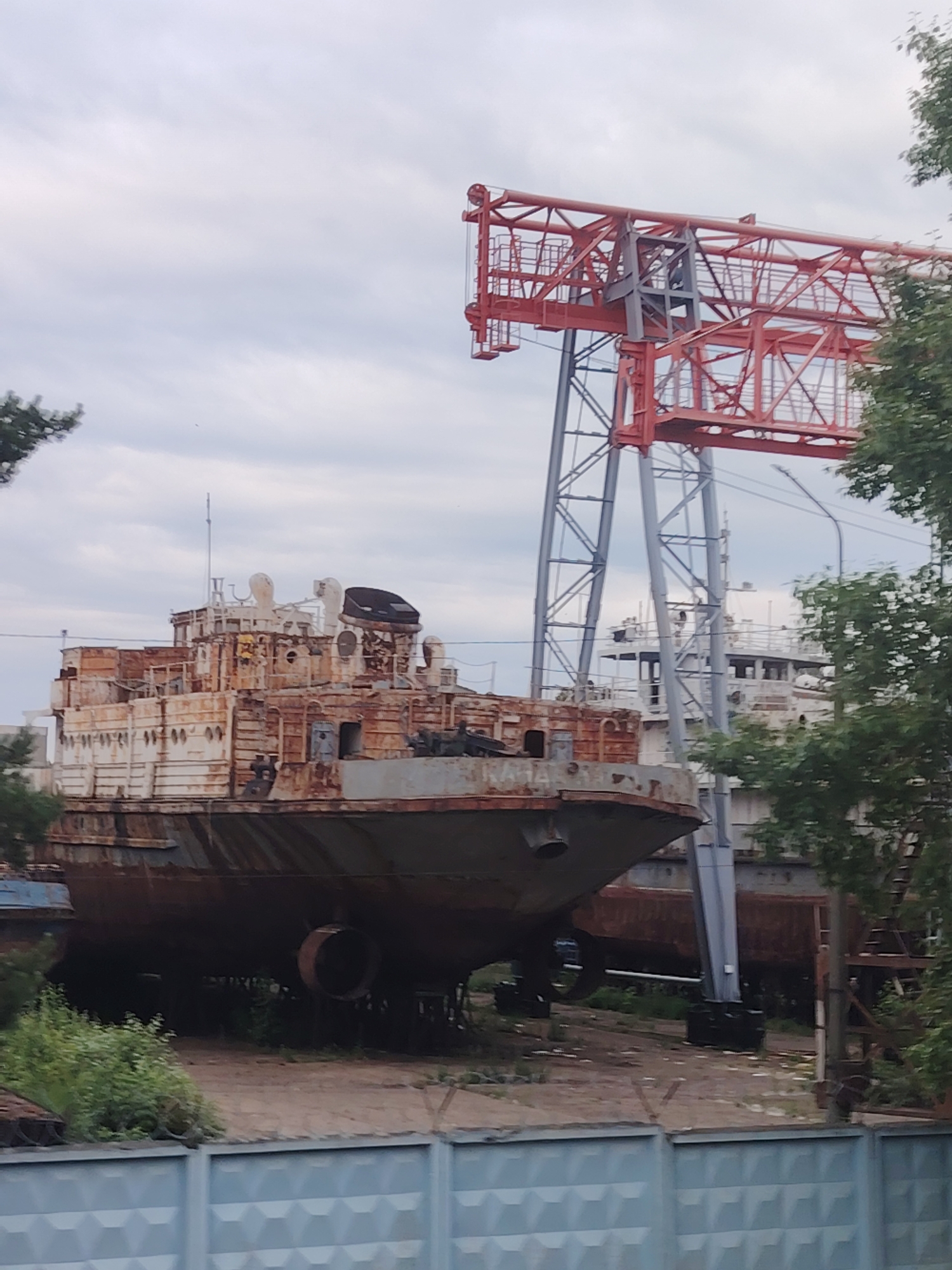 The image depicts a heavily rusted, decommissioned vessel on dry land, likely in a shipyard or industrial area, in the city of Chaikovsky, Russia. The vessel is a large, multi-decked ship with visible signs of deterioration, including peeling paint and exposed metal. A tall, red gantry crane with gray support structures looms in the background, suggesting ongoing or past industrial activity. The sky is overcast. Green foliage from trees is visible on the right and left sides of the frame. A light blue, patterned fence runs along the bottom of the image, separating the viewer from the industrial scene. There are no people visible in the image.