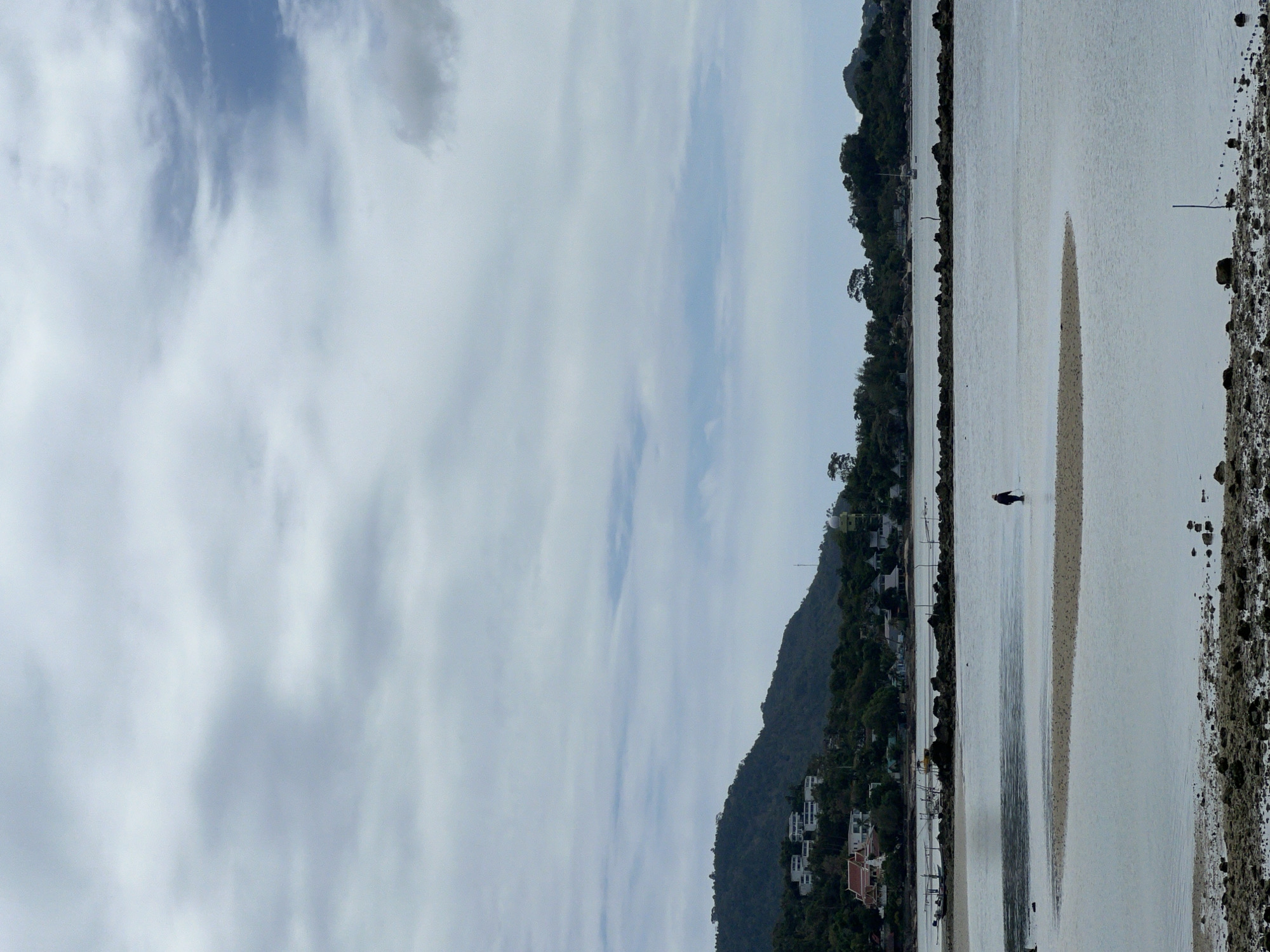 The scene shows a view of the beach at Baan Hua Thanon, Thailand, during low tide. The tide has receded to reveal a wide expanse of sand and shallow water, exposing the seabed. One person is walking in the water. The background reveals a coastline densely vegetated with trees. Buildings, likely residences and resorts, can be seen dotted along the mountainside. The sky is overcast with clouds.