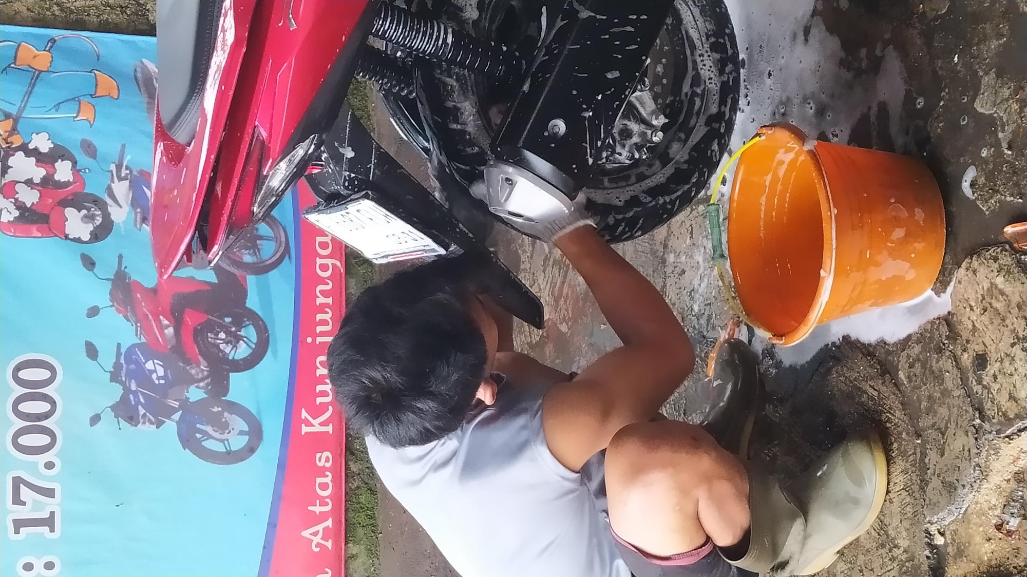 In Cileungsi, Indonesia, a person is washing a red motorcycle. The person, wearing a gray t-shirt, dark shorts, and green rubber boots, is crouched down, cleaning the rear wheel of the motorcycle with a white glove. A bucket of soapy water sits nearby. A banner is visible in the background, with the text "8 17.000" and "Atas Kunjunganga" printed on it, along with images of motorcycles and a person washing a scooter. The ground is wet and appears to be paved with uneven stones.