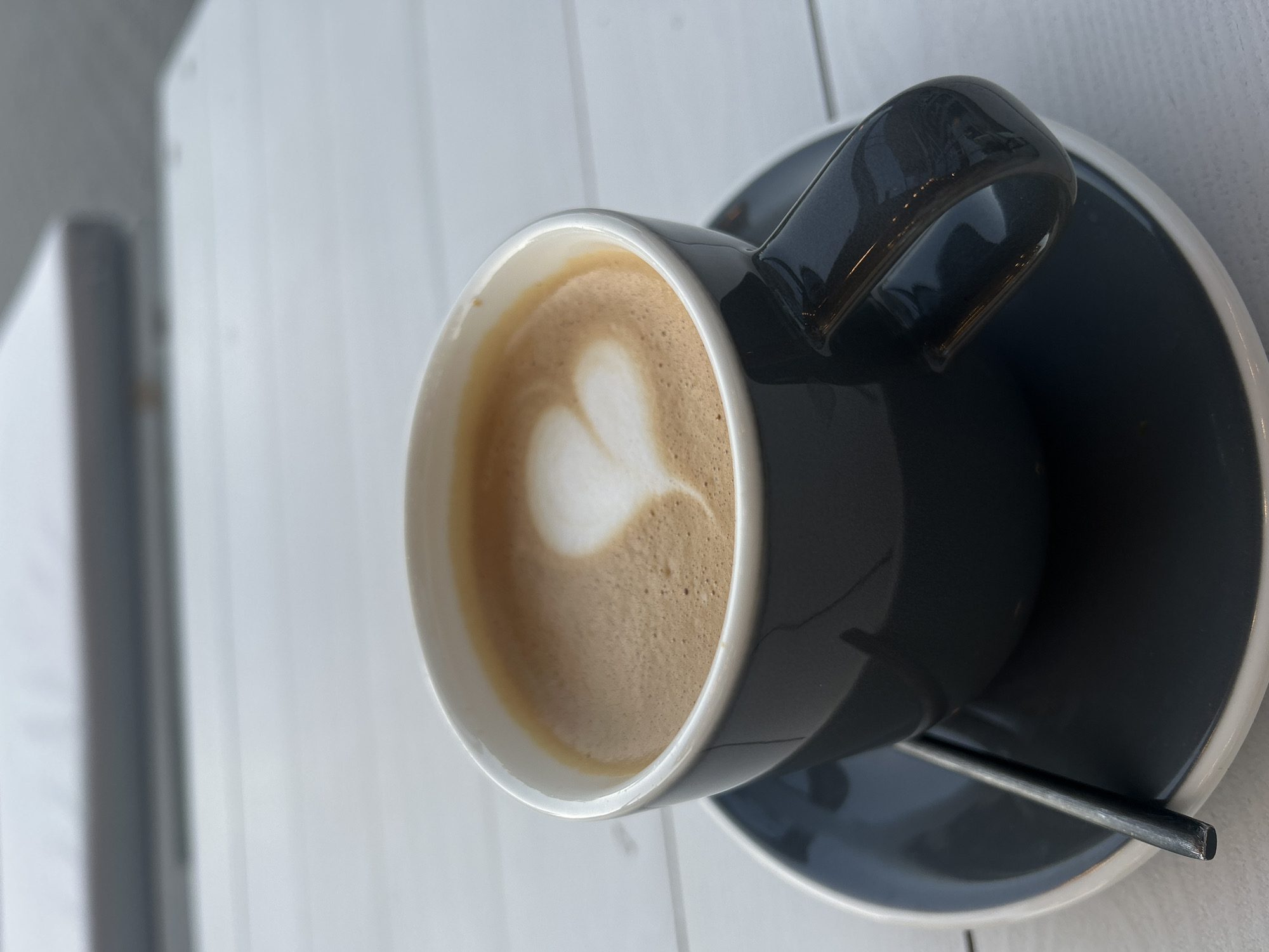 An overhead, slightly angled shot captures a dark gray ceramic coffee cup and matching saucer, set on a light-colored, possibly paneled wooden surface. The cup contains a beverage topped with foamed milk, featuring a distinct, heart-shaped latte art in its center. The coffee itself is a rich brown, transitioning to a lighter, frothy layer of foam. A small, dark gray metal spoon rests on the saucer next to the cup.
The cup has a glossy finish, reflecting some light, particularly noticeable on its handle. The interior of the cup is white, contrasting with the dark gray exterior. The background is softly out of focus, revealing vertical light gray lines against a white or very light gray backdrop, suggesting a wall with panels or blinds. The lighting is bright and even, indicating daytime. The scene evokes a calm and inviting atmosphere, typical of a coffee break. There is no visible text in the image.