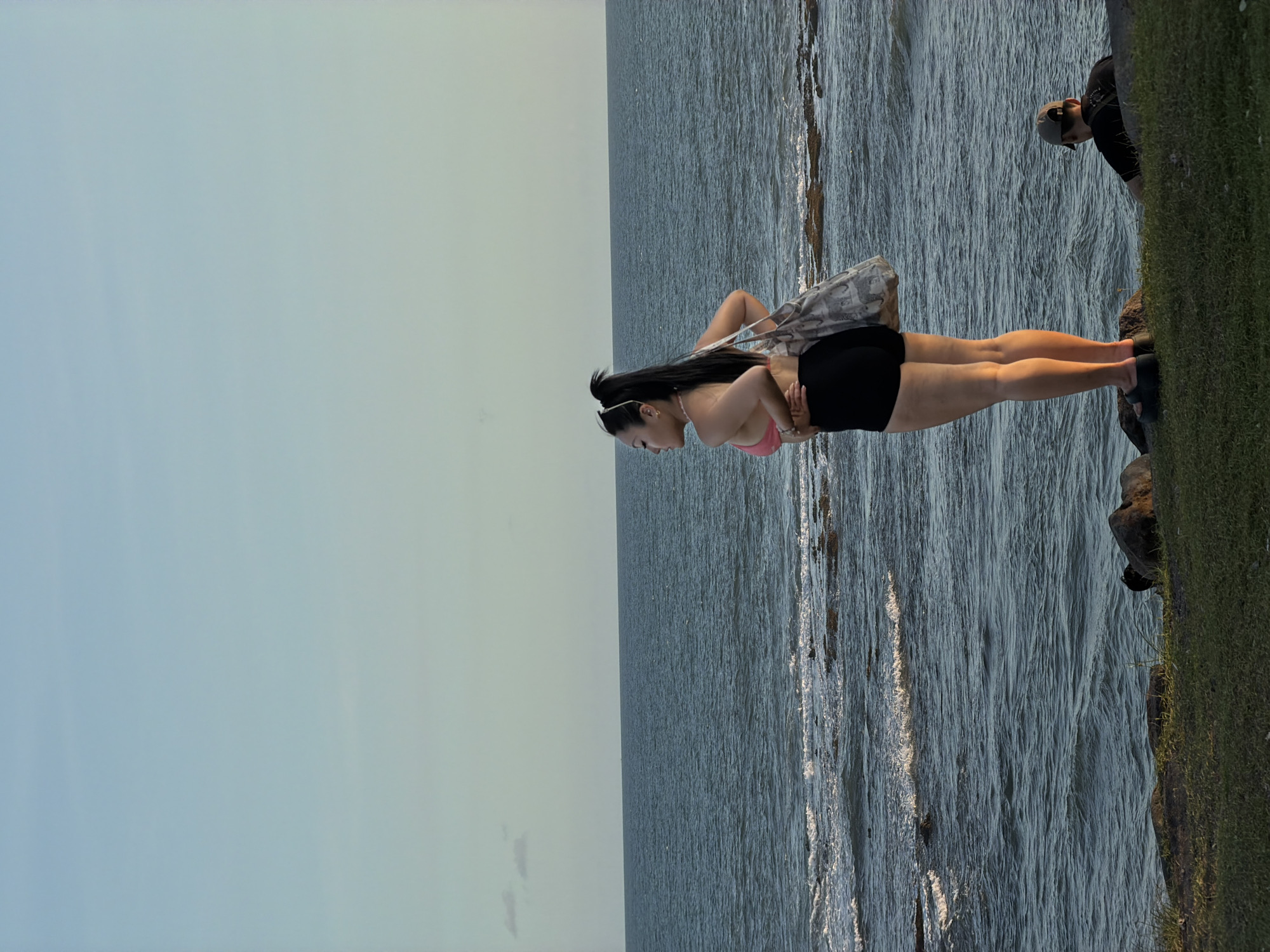 The image captures a scene by the sea in Mengwi, Indonesia. A woman wearing a pink top and black shorts stands on a rocky edge near the water. She is holding a gray tote bag on her shoulder. Behind her, a person with a cap is looking towards her. The sea stretches out in the background, meeting the sky. To the right of the woman is a patch of green grass.