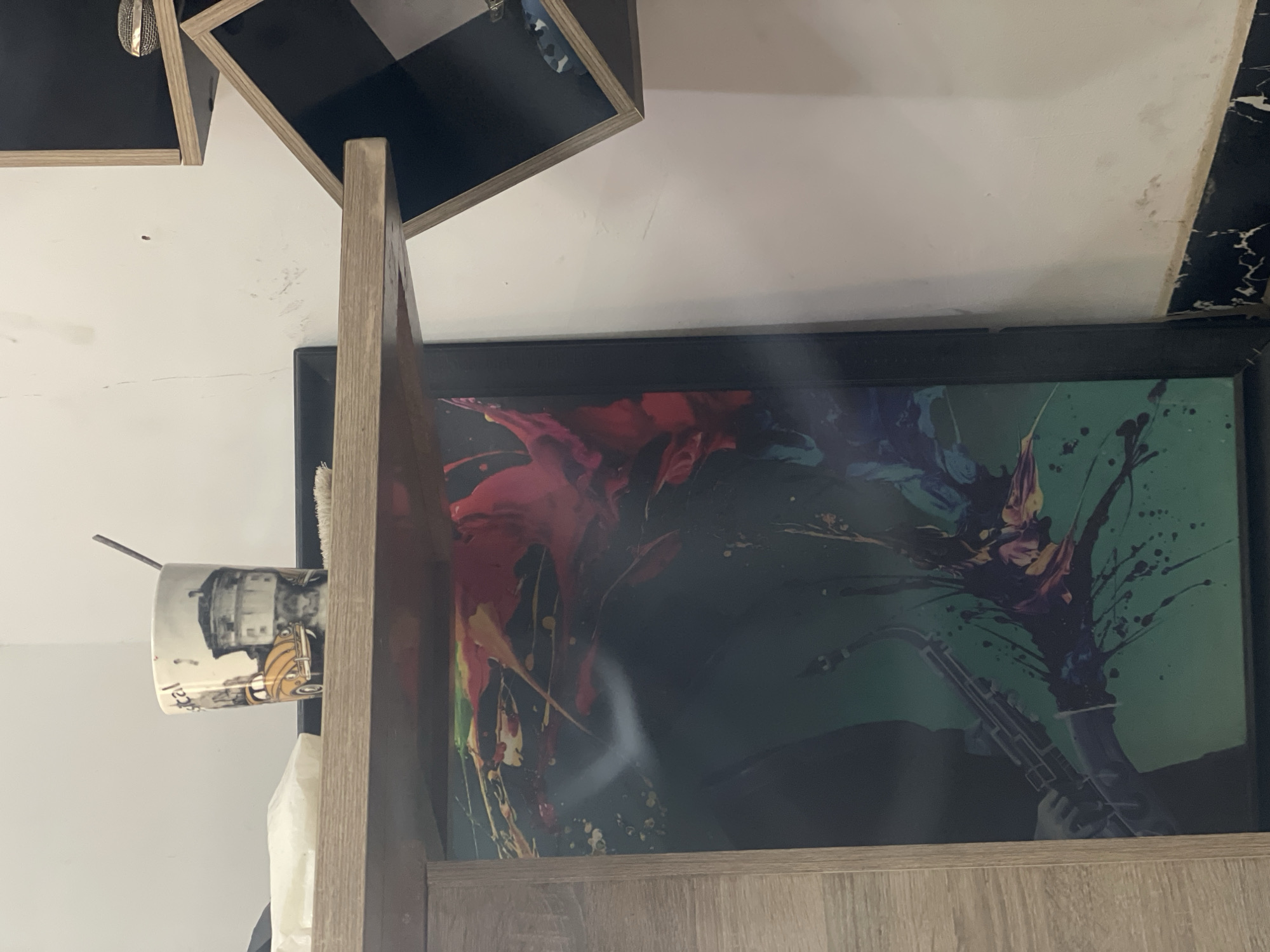 The image displays an indoor scene, likely a wall arrangement, featuring various decorative and functional objects. A prominent light wooden structure, possibly a shelf or part of a larger furniture piece, occupies the center foreground, vertically dividing the view.
To the left of this wooden structure, a white mug is visible, featuring a black and white illustration of a building, possibly a lighthouse or tower, alongside a yellow vintage scooter. A stirring stick or spoon protrudes from the mug. Partially visible, inverted text on the mug spells "Abuja" and what looks like "122" or "221."
Dominating the right side of the image, and partially obscured by the wooden structure, is a large, framed abstract artwork. This piece showcases vibrant splashes of red, pink, yellow, blue, and green paint against a dark background, which transitions from green to black. A faint outline of a saxophone-like musical instrument is discernible towards the bottom right of the artwork.
Above the scene, two square wall-mounted shelves are visible. These shelves have dark, possibly black, interiors and light wooden frames. One shelf contains a small, crumpled metallic mesh object. The wall itself is white, showing some minor marks and textures. In the upper right corner, a piece of furniture or wall trim with a black and white marble-like pattern is partially visible. The scene depicts a static arrangement of items in an interior space, consistent with a room in Abuja, Nigeria.
