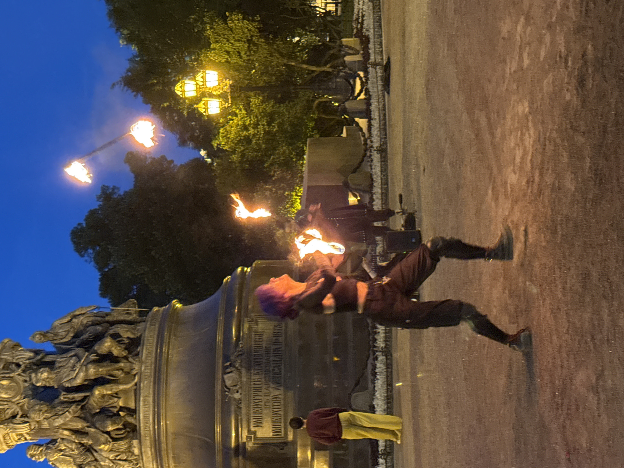 The image depicts a nighttime scene in Saint Petersburg, Russia, in front of the monument to Catherine the Great on Nevsky Prospekt. A fire performer with purple hair is actively juggling torches, with flames visible in the air. The monument, a large bronze sculpture with multiple figures, is illuminated, its inscription legible in the dim light. To the left of the monument, a person stands observing the performance, wearing a maroon sweater and yellow pants. Streetlights cast a warm glow on the surroundings, illuminating trees and the paved ground. The overall atmosphere is one of urban entertainment under a twilight sky.