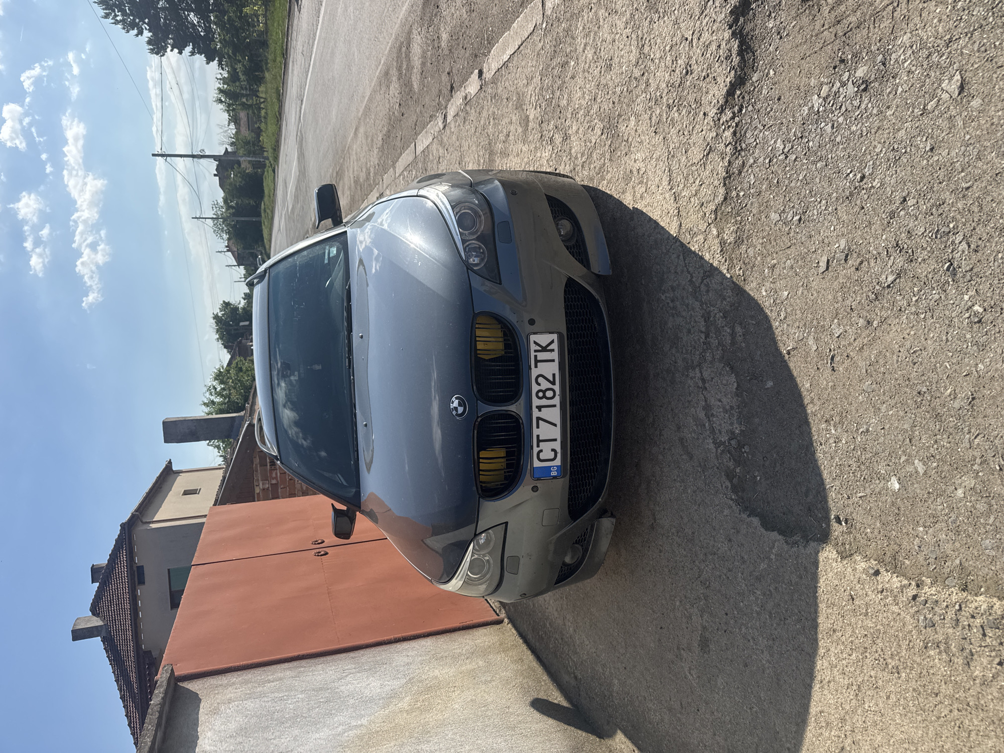 The image shows a gray BMW car with a Bulgarian license plate "CT 7182 TK" parked on a paved surface adjacent to a red-painted building wall. The surroundings appear to be a street in an urban or suburban area, under bright sunlight. The scene is possibly located in or near Sarnevo, Bulgaria, based on the Bulgarian license plate. No people are visible. The image depicts the car in a stationary position, seemingly parked. The environment is characterized by a mixture of buildings and a road, with trees and a blue sky in the background.