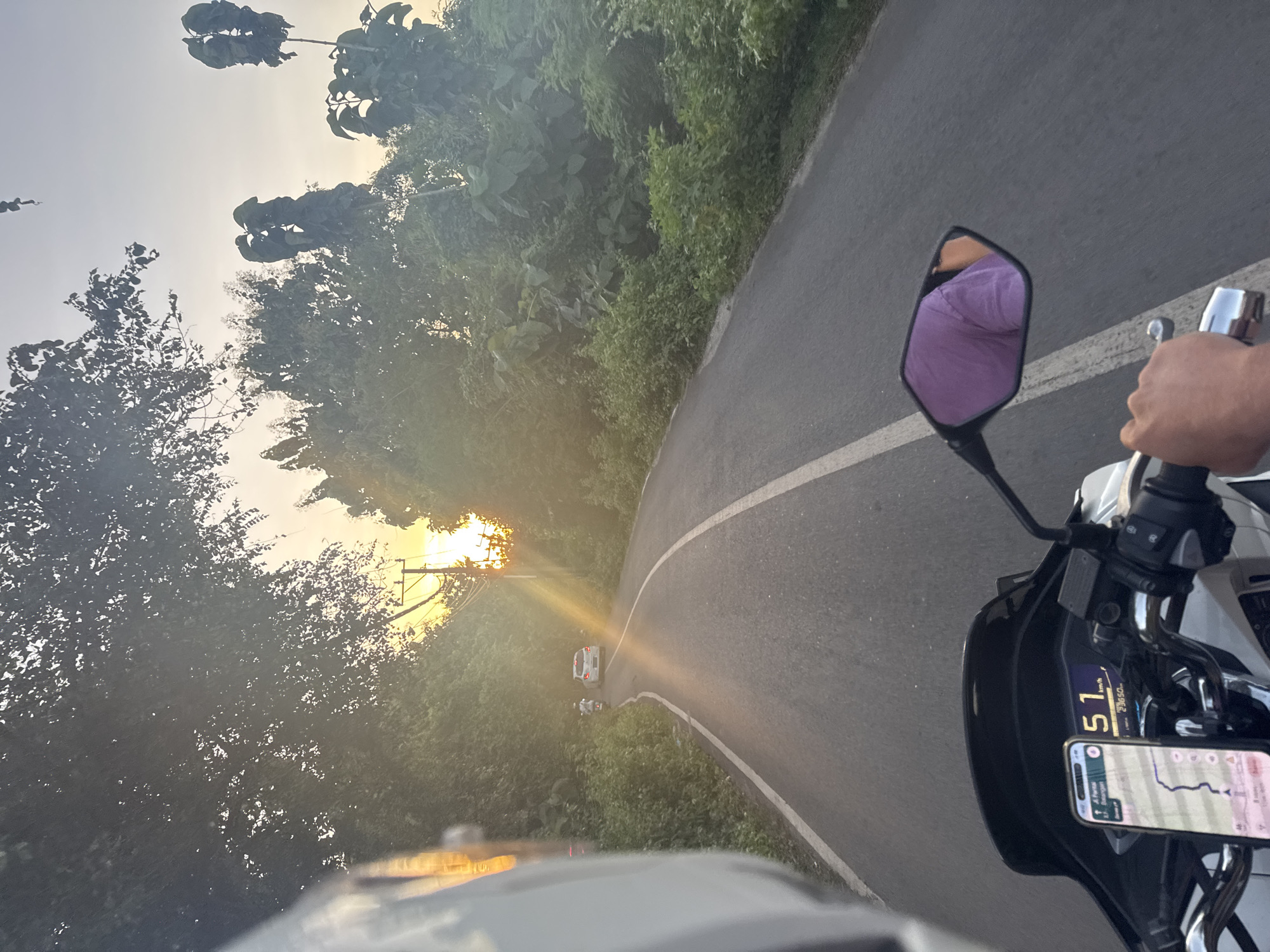 The image captures a point-of-view shot from a motorcycle traveling on an asphalt road. The road curves to the left and is marked with a solid white line. The motorcycle's mirror and a hand gripping the handlebar are visible on the right side of the frame. A smartphone, mounted on the motorcycle, displays a navigation app with a route. The speedometer indicates a speed of 51 km/h. The environment consists of dense green vegetation on both sides of the road. In the distance, there is a car, with the sun setting behind it. The overall atmosphere is bright due to the sunlight. The location is marked on the smartphone as "A, Pertas Balangan."