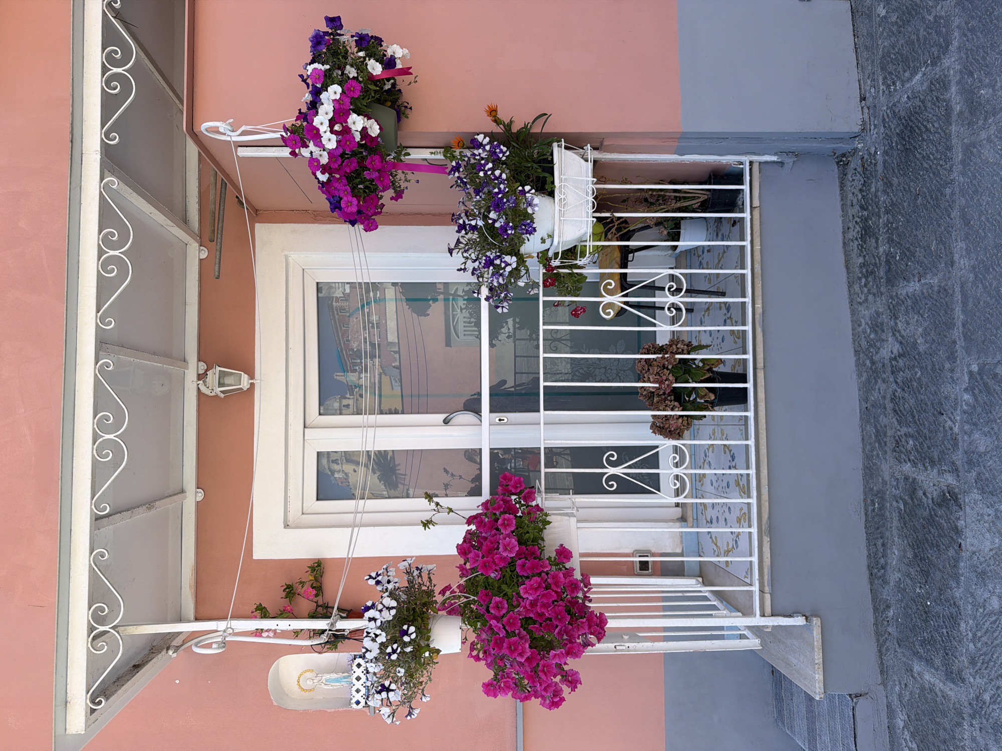 The image depicts a building facade in Procida, Italy. The building is two-toned, with a salmon-pink upper section and a gray lower section. A white-framed window with multiple panes occupies the center of the image. The window reflects buildings and sky. A white wrought-iron railing is positioned in front of the window, adorned with potted plants and hanging baskets filled with vibrant flowers. Three hanging baskets are attached to the front of the window. The first one on the top right side of the image has purple, white and pink flowers. The second is a white pot with purple flowers and marigolds. The last one, on the bottom right side of the image has pink flowers, while the one on the bottom left of the image has white and purple flowers. A small niche containing a statue is visible near the bottom left of the image. A gray stone walkway or street is seen to the right of the building. A white metal awning with decorative swirls extends over part of the window.
