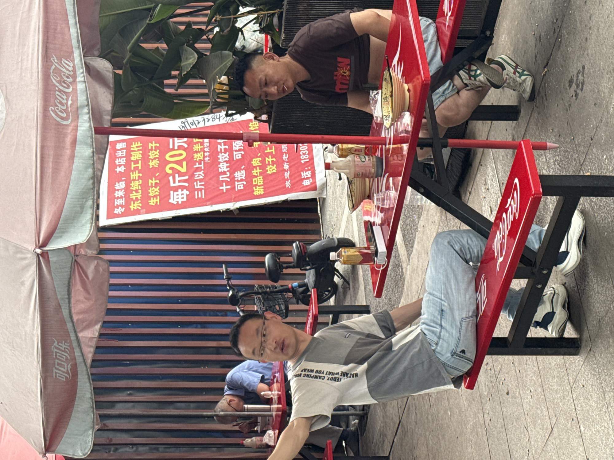 The image shows a street scene in the city of Chongqing High-tech Zone, China, featuring a red Coca-Cola-branded table and chairs set up on a sidewalk. There are three men visible in the image: one man in a brown shirt is seated at the table, looking down. Another man, wearing glasses and a gray and white t-shirt with the words "TEDDY, CAMPING VIRE, HAPARE WHAT YOU WEA," is seated at the table looking up, towards the camera. A third man in a blue shirt is also seated at the table, with his head down. A sign beside the table advertises homemade northeastern-style dumplings for 20 yuan per kilogram. There's a bicycle parked behind the table. A Coca-Cola-branded umbrella is visible overhead. The environment appears to be urban, with a striped blue and red backdrop behind the table. There are bottles and bowls on the table.