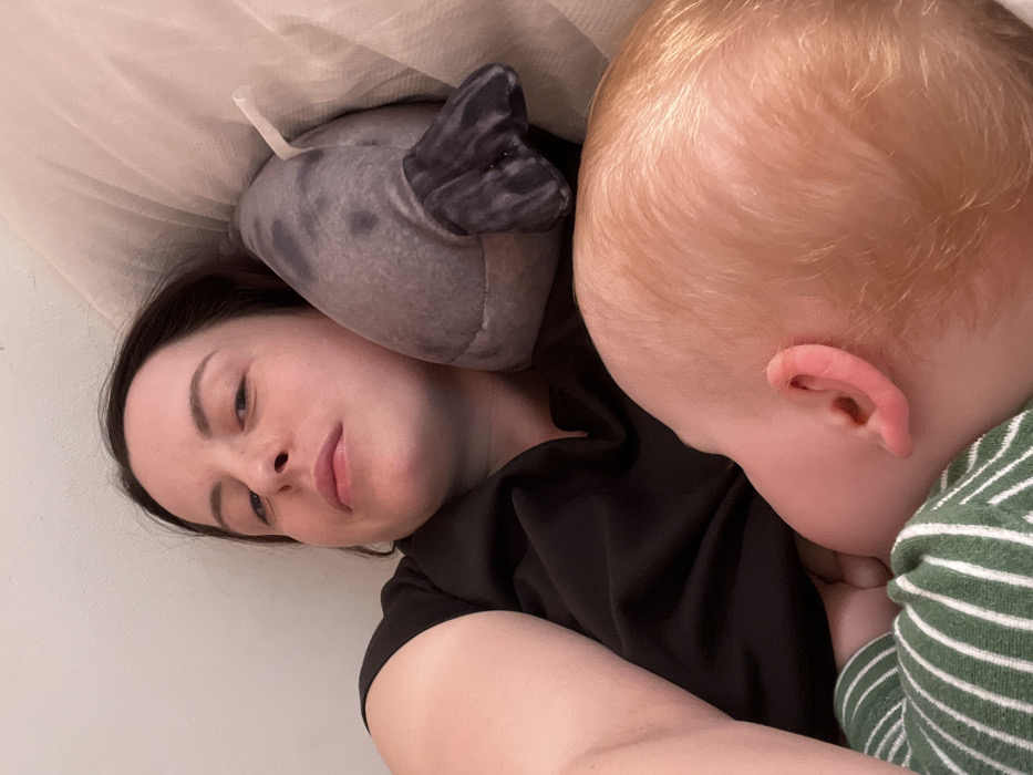 The scene depicts an adult female and an infant in an indoor setting within Hohenbrunn, Germany. The adult female, with dark hair and a neutral facial expression, is lying on her back, looking directly towards the camera. She wears a black short-sleeved top. An infant with light, possibly red or blonde, hair is positioned beside her, actively feeding from her chest. The infant's head and upper torso are visible, and it is dressed in a green and white striped garment. A grey plush toy, resembling a seal or manatee with darker flippers and a white tag, rests near the adult female's head/shoulder. The immediate environment consists of light-colored bedding or pillows, suggesting a bed or couch. The background is a plain, light-colored surface.