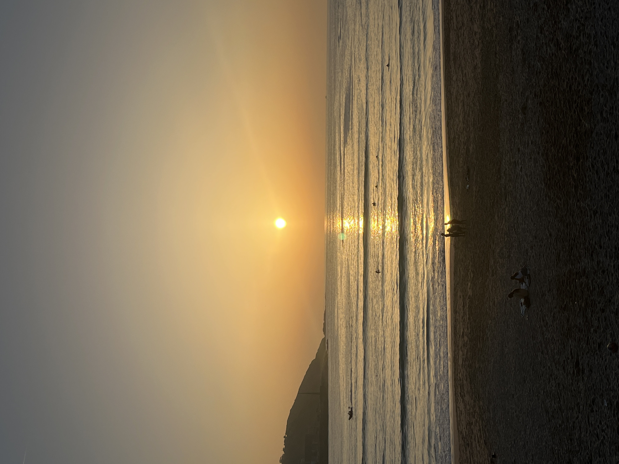 The image depicts a sunset over the ocean, with the sun positioned near the horizon. Sunlight reflects on the surface of the water, creating a bright line. A beach borders the water, appearing darker in tone, and shows the silhouettes of a group of people. The sky exhibits a gradient of color, transitioning from bright yellow near the sun to a muted gray. A dark landmass is visible along the horizon. The location is A Coruña, Spain, given the presence of the cityscape.