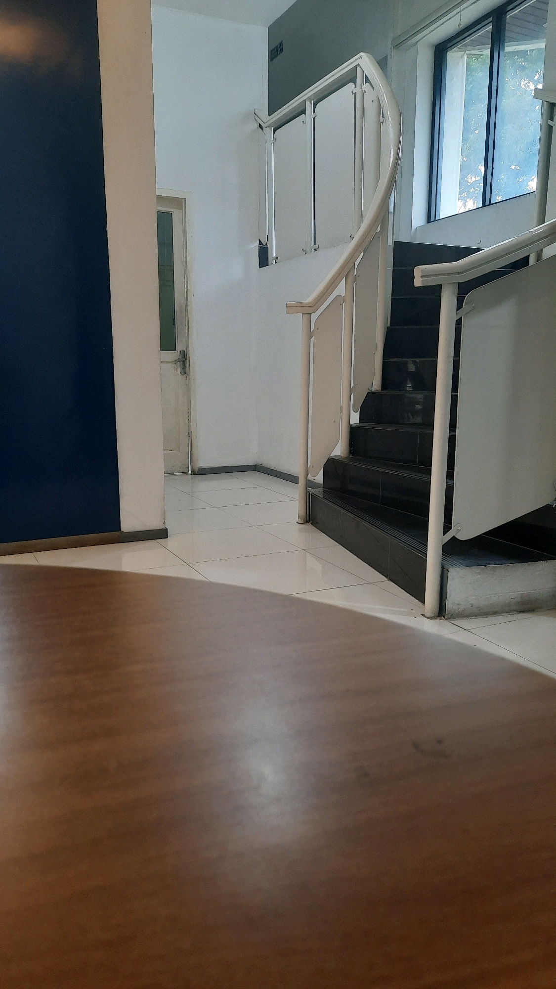 The image shows an interior view of a building's staircase and hallway, likely in Padang, Indonesia, given the context of the request. In the foreground, a portion of a polished wooden surface, possibly a table or floor, is visible, out of focus. Moving towards the background, a hallway with white tiled flooring leads to a white door with a metal handle and a small window pane. To the left, a portion of a dark blue wall is present. Adjacent to the hallway, a white staircase with a curved white railing ascends. The stairs themselves are dark, appearing to be made of black stone or tile. The walls surrounding the staircase are painted white, and a window with a view of trees and sky is visible at the top of the stairs. A discreet green sign with white text is mounted on the wall above the staircase. The lighting suggests an indoor environment, likely during daylight hours. There are no people or specific actions depicted in the scene.