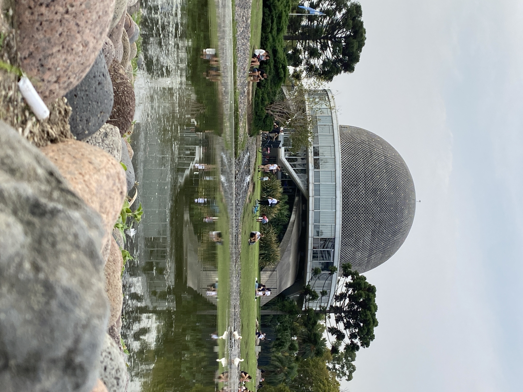 The image depicts the Galileo Galilei Planetarium in Buenos Aires, Argentina, reflected in a body of water, likely a lake or pond. The foreground is dominated by large, rounded stones. A discarded cigarette butt is visible among the stones. Beyond the stones, water reflects the planetarium and surrounding greenery. People are visible on the lawn adjacent to the building, some standing, others sitting. Trees are interspersed throughout the scene, including prominent pine trees. The sky is overcast.
