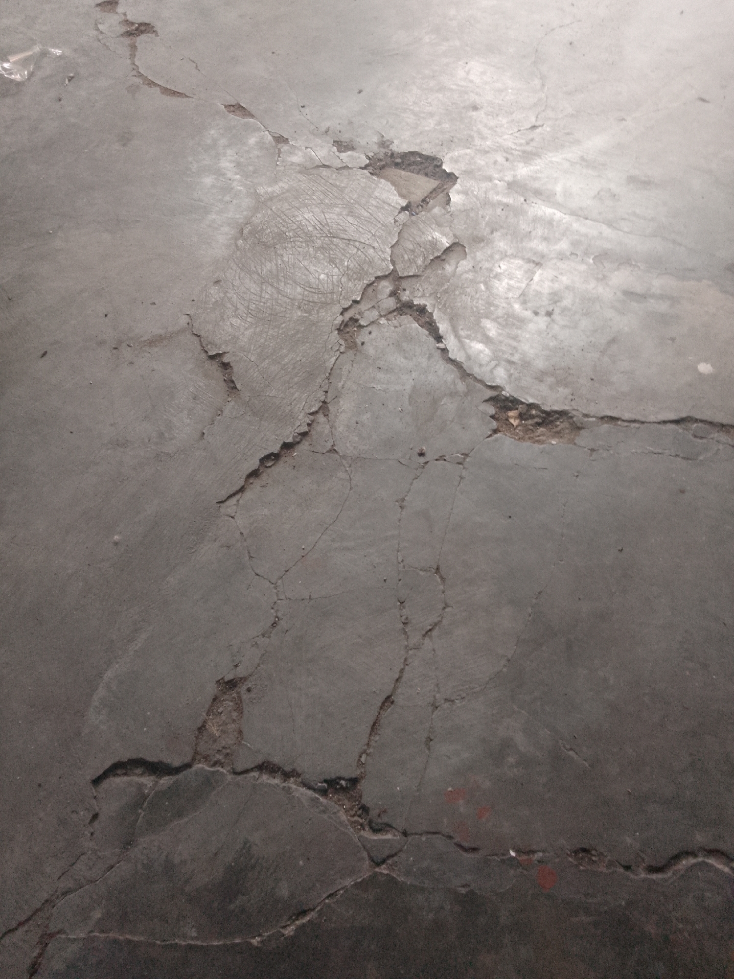A close-up, angled shot depicts a damaged concrete surface. The concrete is grey and exhibits numerous cracks, some of which are wide and reveal the underlying material. There are also areas where the concrete has chipped away, creating rough, uneven patches. In some sections, faint circular markings or scratches are visible on the surface, possibly from tools or equipment. A small piece of translucent plastic is visible in the upper left corner. The lighting creates subtle shadows and highlights, emphasizing the texture and imperfections of the concrete. The scene appears to be an outdoor environment, possibly a pathway or floor. The image does not contain any people or animals. The location in the city of Trenggalek, Indonesia, is not discernible from the visual information provided in the image.