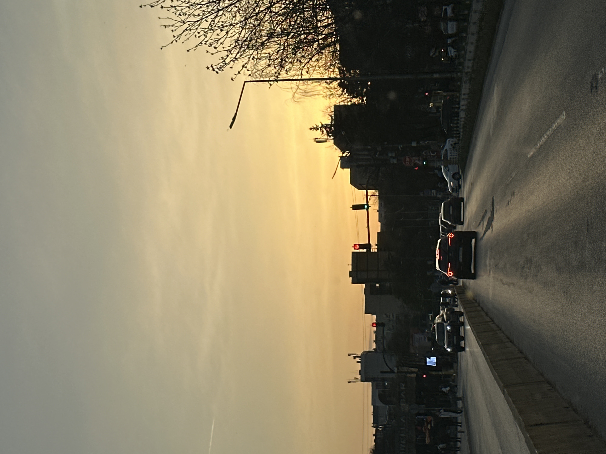 The image depicts a street scene in Sofia, Bulgaria, at sunset. The sky is a gradient of yellow and grey. A road runs along the right side of the frame, with several cars driving on it. A row of buildings and trees lines the left side of the road. Traffic lights are visible along the street.