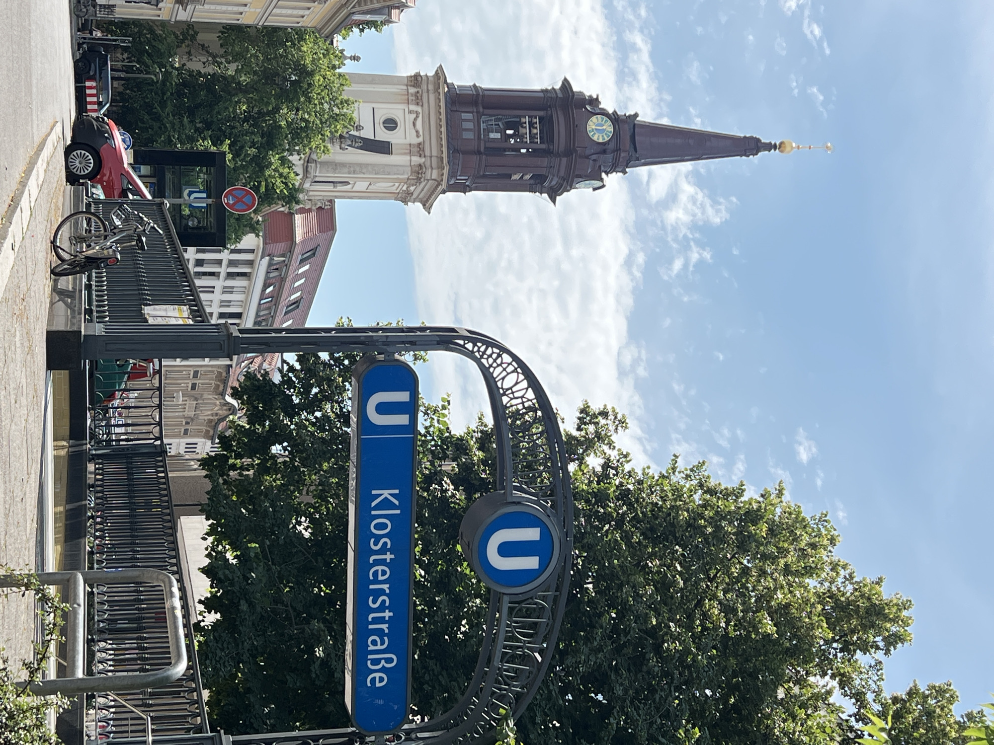 This is a daylight, eye-level, outdoor, medium shot of the U-Bahn station entrance at Klosterstraße in Berlin, Germany. On the right side of the frame, a blue U-Bahn sign with the station name "Klosterstraße" is displayed on an ornate metal archway. Behind the sign, lush green trees form a dense canopy. To the left, a tall, historic church tower with a clock face stands prominently against a bright blue sky with scattered white clouds. At the base of the tower, buildings with red-tiled roofs are visible. In the foreground, a sidewalk runs alongside a street. A red van and a black SUV are parked on the street. A bicycle is parked on the sidewalk near the U-Bahn entrance. A circular sign with a red border and a horizontal bar across a red circle, indicating no stopping, is visible near the subway entrance.