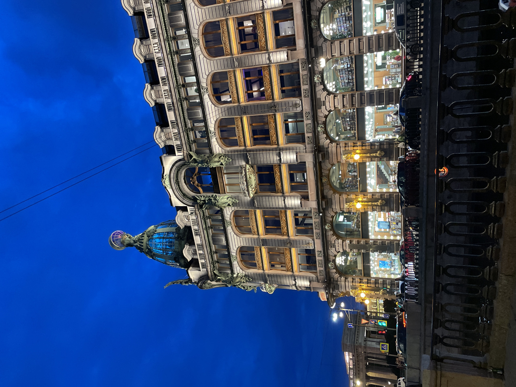 A grand, ornate building stands prominently against a deep blue twilight sky, its illuminated windows casting a warm glow. The architecture features intricate detailing, arched windows, and a striking dome topped with a globe and figure. On the street in front of the building, cars are parked and driving, and several pedestrians are visible. A dark metal railing runs along a canal or river in the foreground, separating it from the street and building. The scene suggests an evening in a vibrant urban setting, likely a historic city center. The location is identified as the Dom Knigi (House of Books) on Nevsky Prospekt in Saint Petersburg, Russia.