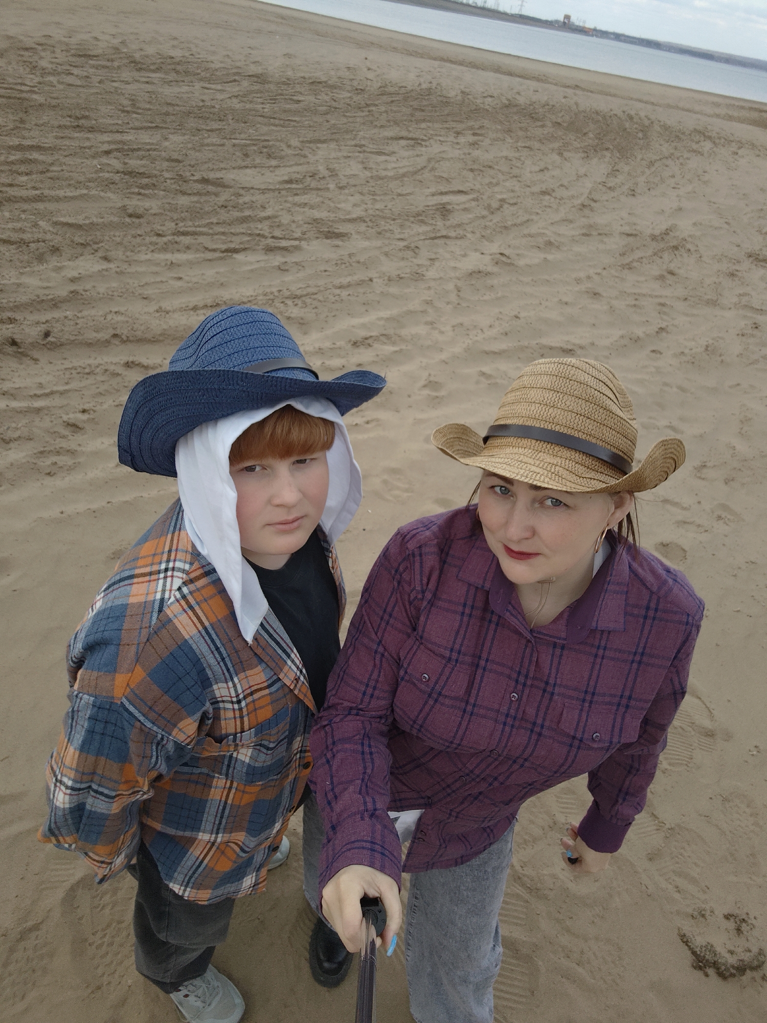 Two individuals are captured in a high-angle shot on what appears to be a sandy beach. The individual on the left is wearing a blue cowboy hat over a white head covering, a blue and orange plaid shirt, dark pants, and grey shoes. The person on the right is wearing a tan cowboy hat with a brown band, a purple plaid shirt, and grey jeans. The person on the right holds a selfie stick in their hand. The background features a sandy expanse leading to a body of water and a distant industrial landscape. The city of Noviy, Russia, is not discernible from the visual information provided.