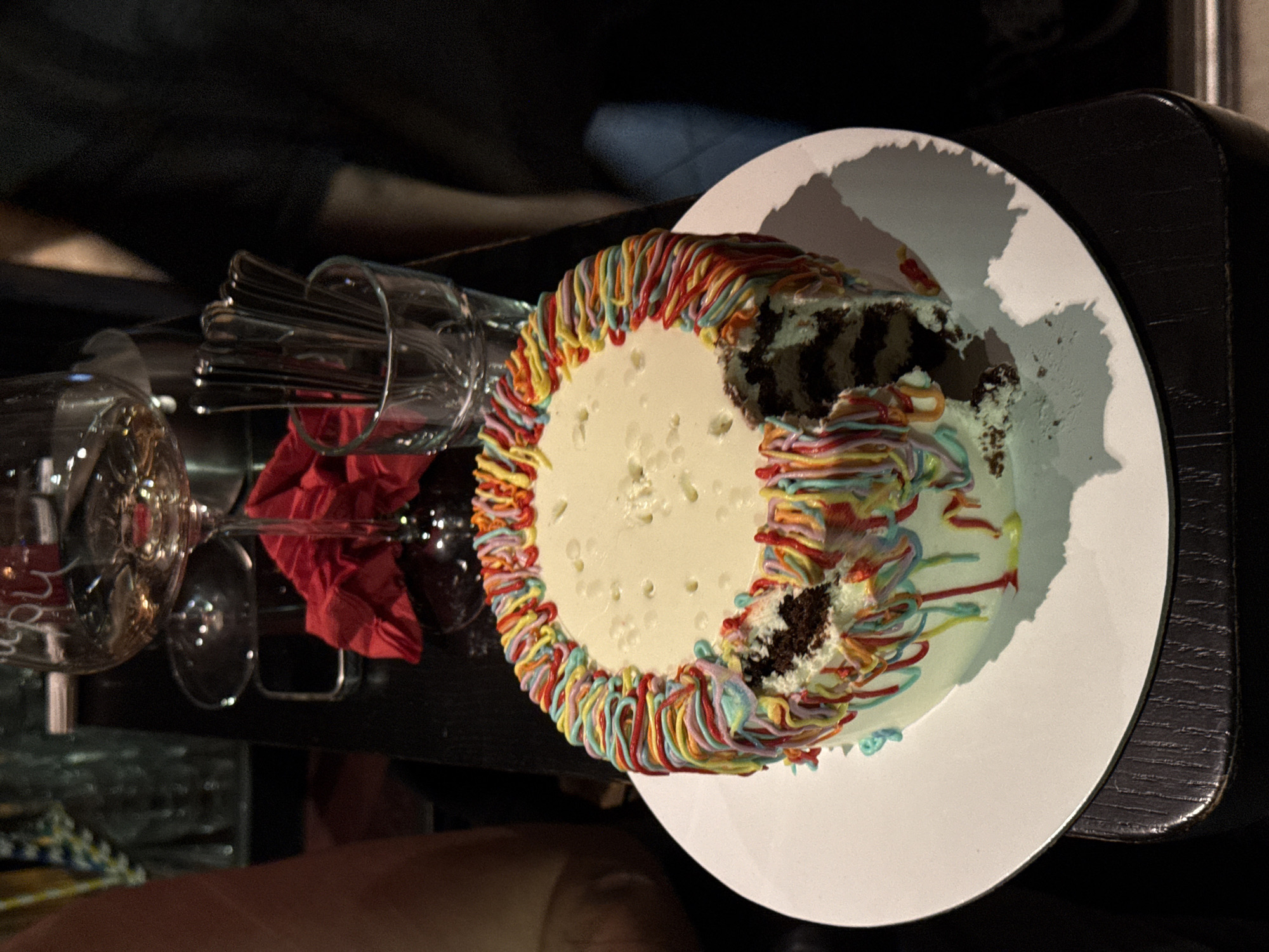 The image captures a partially eaten cake on a white plate, situated on a dark, possibly wooden, surface. The cake is adorned with colorful icing around the edges and has a white top layer, with noticeable missing slices revealing a dark cake interior. To the left of the cake, there are wine glasses and a container holding cutlery, along with a red napkin. The image appears to be taken indoors, potentially in a restaurant or bar, possibly in the city of Belgrade, Serbia, though no specific location is identifiable in the image. The overall lighting is subdued.