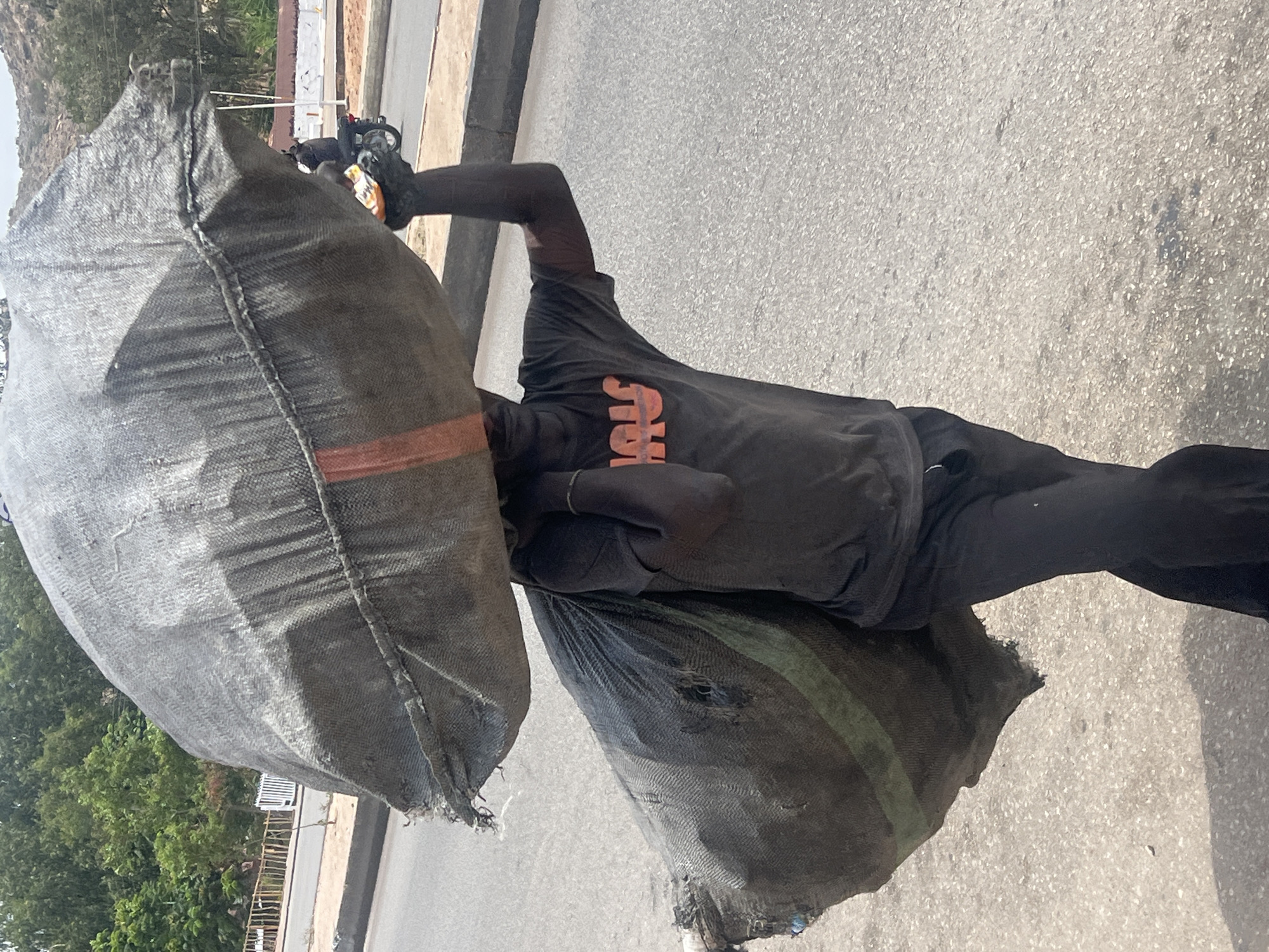 The image, taken in Jos, Nigeria, captures a person carrying two substantial, dark-colored woven bags on a sunny day. The individual, likely male, is centrally positioned, walking on what appears to be an asphalt road. He wears a dark short-sleeved t-shirt with orange letters "SIM" visible on the chest, and dark long pants.
One large, possibly burlap-like bag rests on his left shoulder, largely obscuring his head and upper body. A second, similarly worn bag, distinguished by a green stripe, is carried lower on his right side. Both sacks show signs of wear and tear, with visible fraying and small holes, suggesting they are heavily used for transporting goods. The person's posture conveys the effort involved in carrying the heavy load.
The setting is an outdoor street or pathway, bathed in bright sunlight that casts distinct shadows. In the background, lush green trees and what appears to be a distant hillside or mountain are visible on the left. On the right, hints of a light-colored structure and a parked motorcycle or scooter can be seen. The road surface is light grey, characteristic of asphalt. The scene depicts a common activity of labor or transport in a warm, bright climate.