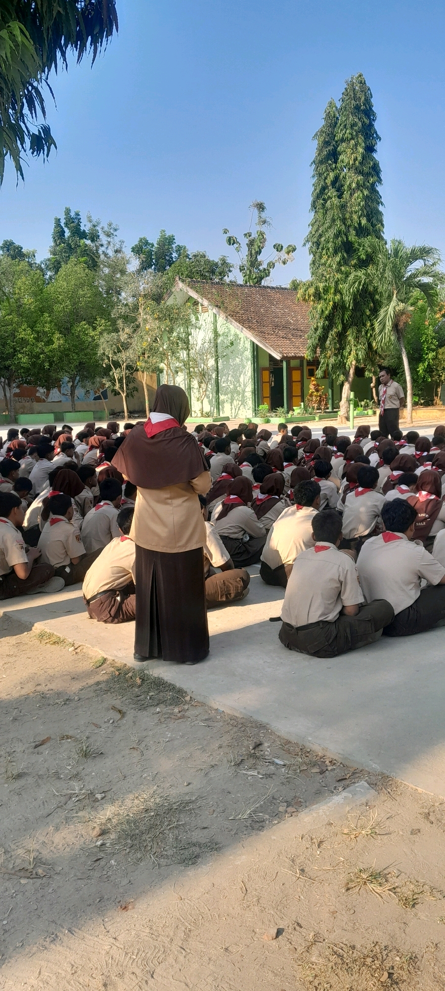 In the city of Tahunan, Indonesia, a group of young people, likely students from a scouting organization, are seated on a paved area in front of a light green building with a brown tiled roof. The students are wearing tan uniforms with red and white neckerchiefs. One adult, also in uniform and wearing a hijab, stands facing the seated group, appearing to address them. Another adult male, in uniform, stands to the right, also facing the students. The environment is outdoors, with trees and foliage visible in the background, and the sky is a clear blue. The ground is a mix of paved surfaces and dirt with sparse vegetation. The lighting suggests it is daytime, with sunlight casting shadows.