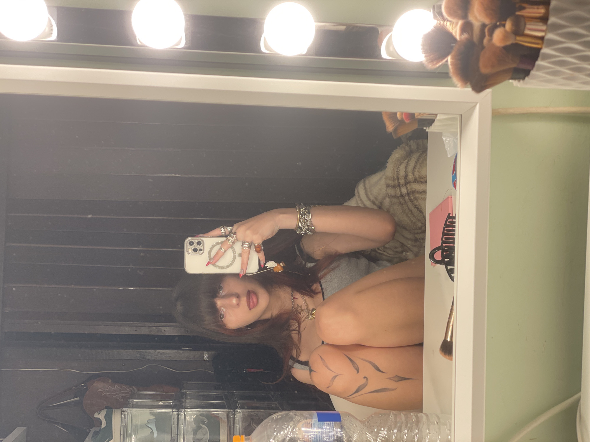 The image presents a mirror selfie of a young female in an indoor setting, consistent with a private room or dressing area within Petah Tikva, Israel. The subject, positioned centrally, is reflected from the waist up, facing forward with her tongue slightly extended. She has dark hair with bangs and lighter reddish streaks, and wears a gray tank top. Her right hand holds a light-colored smartphone, likely an iPhone, equipped with a magnetic ring holder and a dangling charm, directed towards the mirror to capture the reflection. Multiple rings adorn her fingers, and a collection of bracelets is visible on her left wrist. A black, stylized tattoo featuring a heart-like design with radiating lines is prominently displayed on her right thigh, which is bent.
The selfie is taken in front of a white-framed vanity mirror, illuminated by several visible round light bulbs arranged vertically on its right side. On the white surface directly below the mirror, a makeup brush with a gold handle and a black claw hair clip are present. To the far left, a clear plastic water bottle is partially visible. The reflection in the mirror behind the subject reveals dark, vertically paneled walls. On the left side of the reflection, clear plastic stackable storage containers, possibly holding shoes, are visible, along with a brown handbag hanging above them. A light-colored, patterned blanket or fabric is reflected behind the subject's right arm.