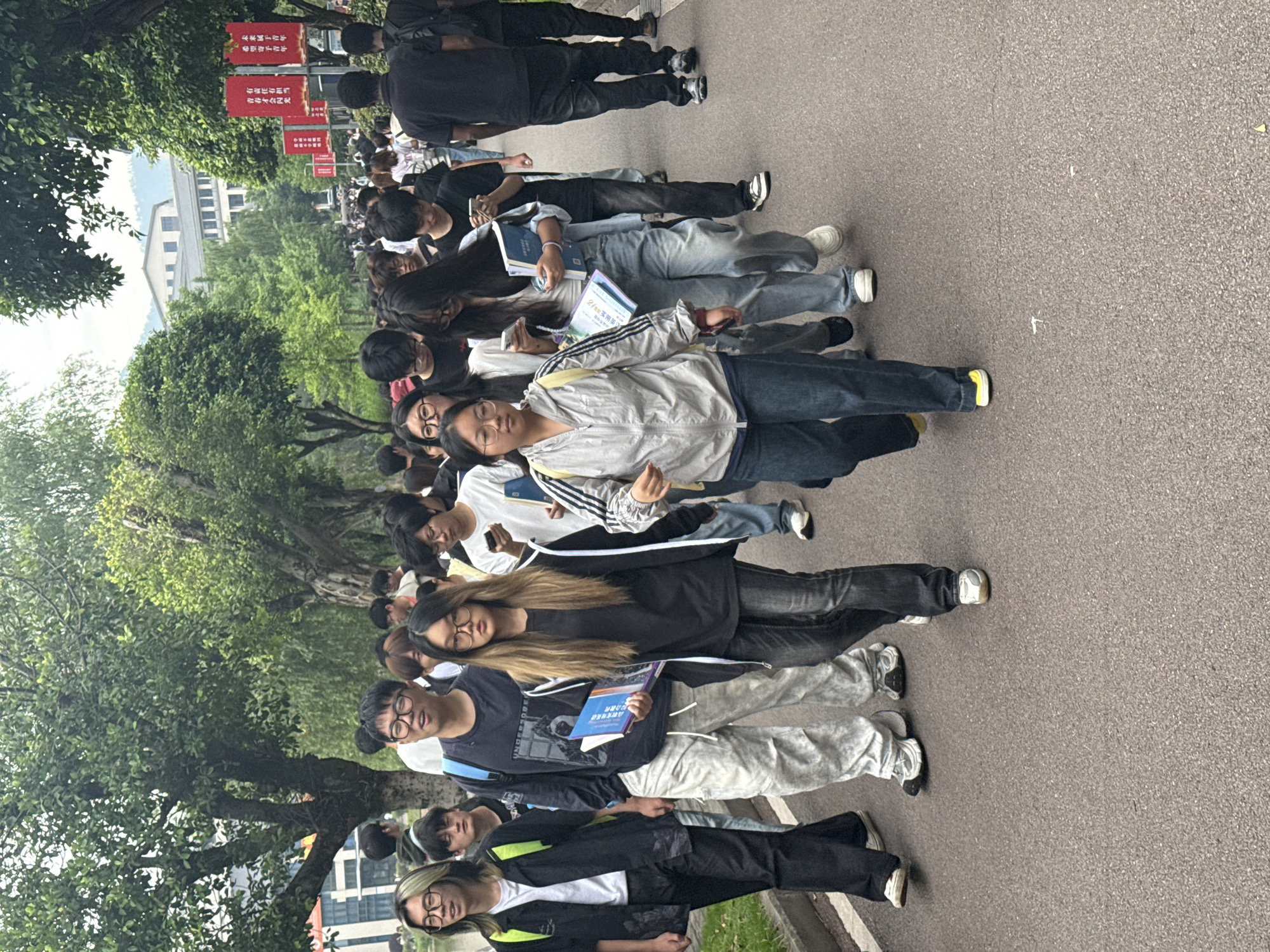 The image shows a line of people standing on a paved road, likely a street, with trees lining one side. The people appear to be young adults, possibly students, as some are holding books. The atmosphere is likely outdoors on a cloudy day. Red signs with Chinese characters can be seen in the background. Based on the provided data, this is in Jiangjin, China.