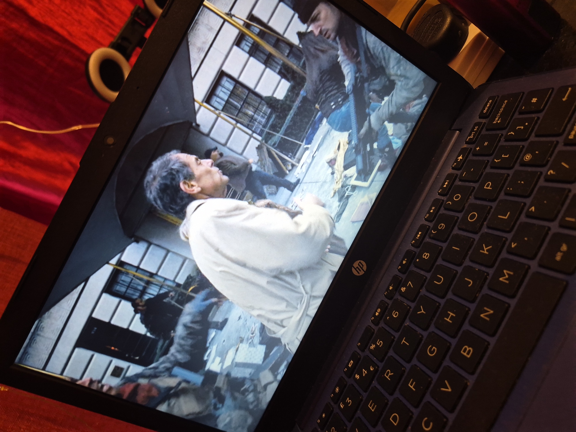 An overhead shot displays a laptop open and angled to the viewer, with a scene from a film or television show on its screen. The on-screen image depicts a street scene with several individuals. In the foreground, a man in a light-colored jacket is visible in profile, looking down and to the right. Behind him, another man in a dark jacket and blue jeans walks away from the camera. Further back, a third man in a dark coat is partially obscured, and a fourth individual in a dark outfit appears to be holding a rifle. The background shows a building with multiple windows. The laptop's keyboard, in dark blue with yellow lettering, is partially visible in the lower portion of the image. The laptop is situated on a textured, red surface. A thin yellow wire hangs from the top right of the frame, and a black speaker with a circular white element is visible above the laptop. The scene is captured in a way that suggests it is taking place at night or in low light conditions, and there are no visual cues to indicate it is specifically located in Sandwell, United Kingdom.