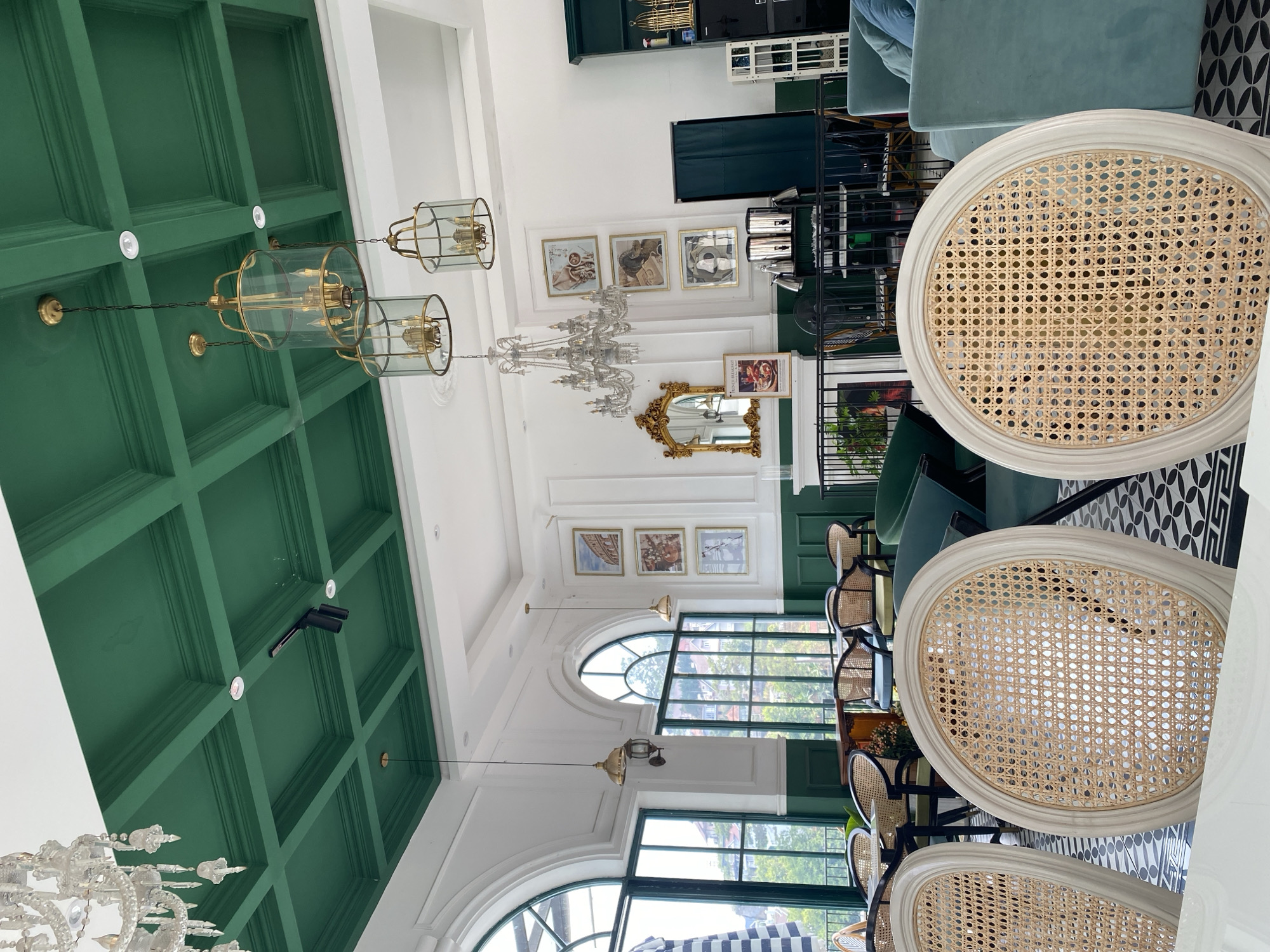 This image captures the elegant interior of a restaurant or cafe, likely in Da Lat, Vietnam, characterized by a refined and classic design. The space features a striking emerald green coffered ceiling that contrasts sharply with pristine white walls and additional white ceiling sections.
Various seating arrangements are visible, including several white-framed chairs with natural rattan cane webbing on their backs and seats, suggesting a colonial or French bistro aesthetic. Towards the back and left, plush light blue or teal velvet sofas and armchairs offer a softer seating option. The floor is adorned with a decorative black and white geometric tiled pattern.
Lighting is provided by a mix of natural light streaming through large arched windows, which reveal glimpses of an outdoor urban landscape, and artificial fixtures. Ornate crystal chandeliers hang from the white ceiling, while brass and glass pendant lights are suspended from the green coffered ceiling. Recessed spotlights are also integrated into the ceiling design.
The white walls are decorated with several framed artworks, predominantly with golden frames. A prominent feature is an ornate gold-framed mirror. Next to it, a framed print depicting a woman is visible, with the text "FRENCH BREAKFAST" clearly legible below. Other framed pictures show still life scenes or architectural details. The overall ambiance is bright, sophisticated, and inviting, suggesting a daytime setting. No people are visible in the scene.