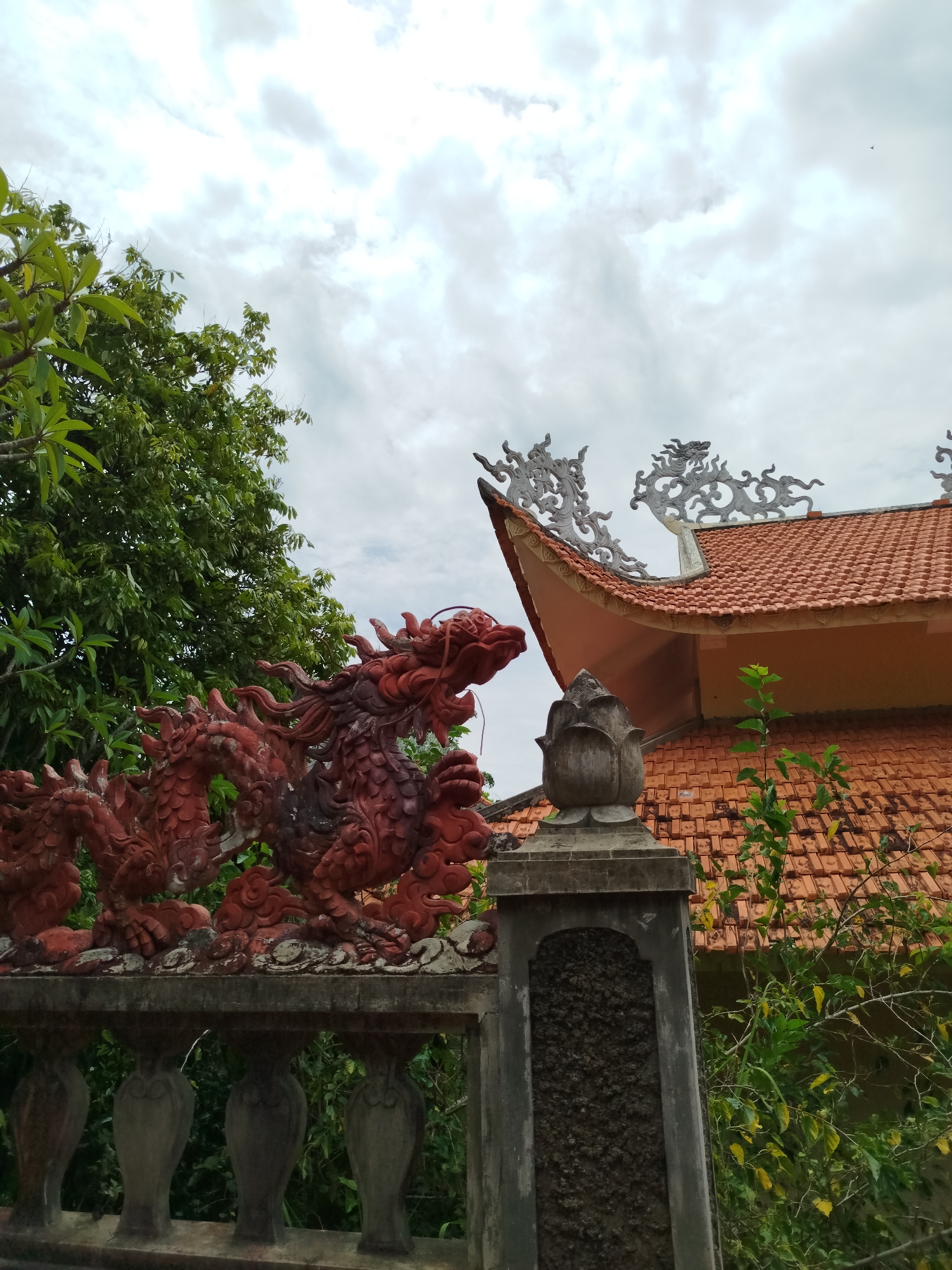 A red dragon statue is perched atop a concrete fence in Da Lat, Vietnam. The fence surrounds a building with an orange tiled roof adorned with ornamental white carvings. Green trees are visible behind the dragon and the building. The sky is overcast with white clouds.