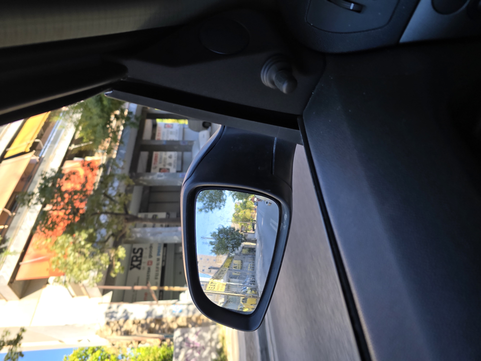 The image, captured from inside a vehicle, shows a view through the side mirror and the passenger side window. The side mirror reflects a street scene in Athens, Greece. In the reflection, yellow taxis are parked along the side of a street lined with trees and buildings. One building in the background has the text "XRS" visible on its facade. The passenger side window shows a blurred view of a building and trees outside, consistent with a moving vehicle. The interior of the car, including the car door and mirror housing, is visible in the foreground. The lighting suggests it is daytime, with sunlight illuminating the street scene.